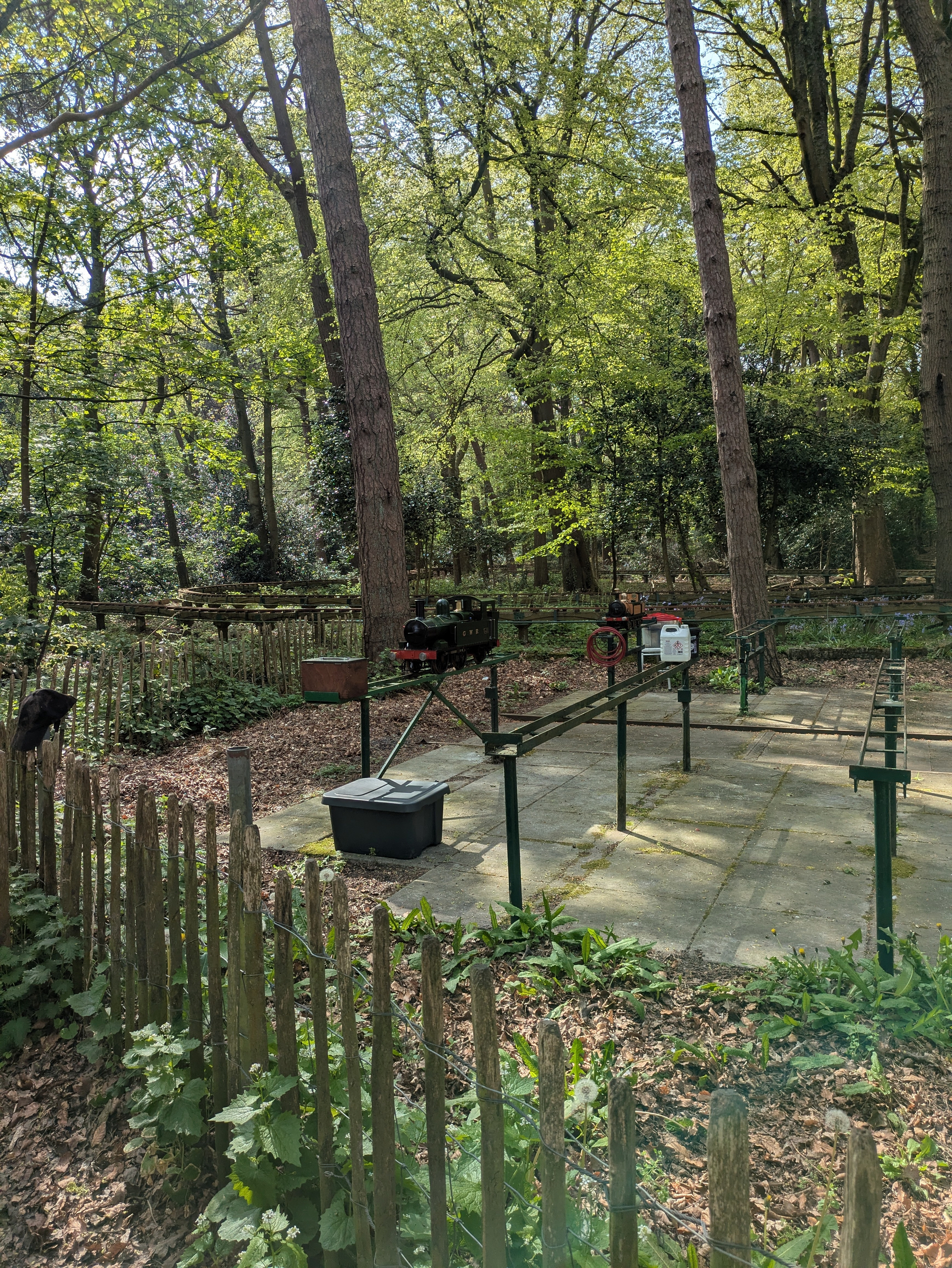 A small train track with a miniature train set is surrounded by lush green trees and a wooden fence in a forested area.
