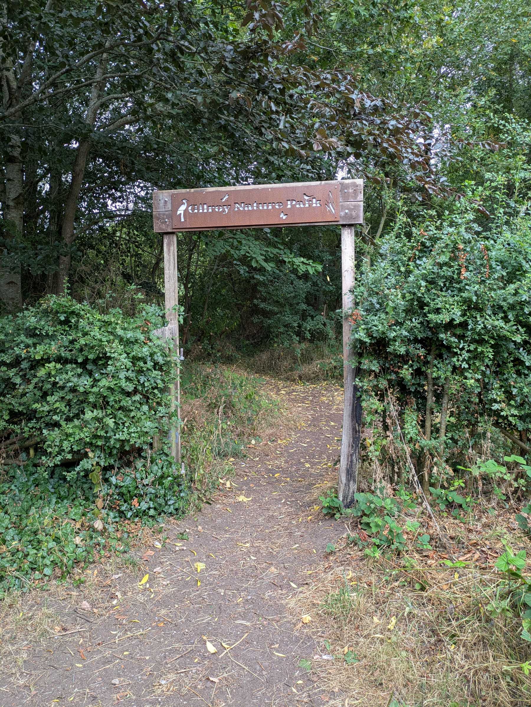 A dirt path leads through an entrance marked by a wooden sign for Gilroy Nature Park, surrounded by dense green foliage.