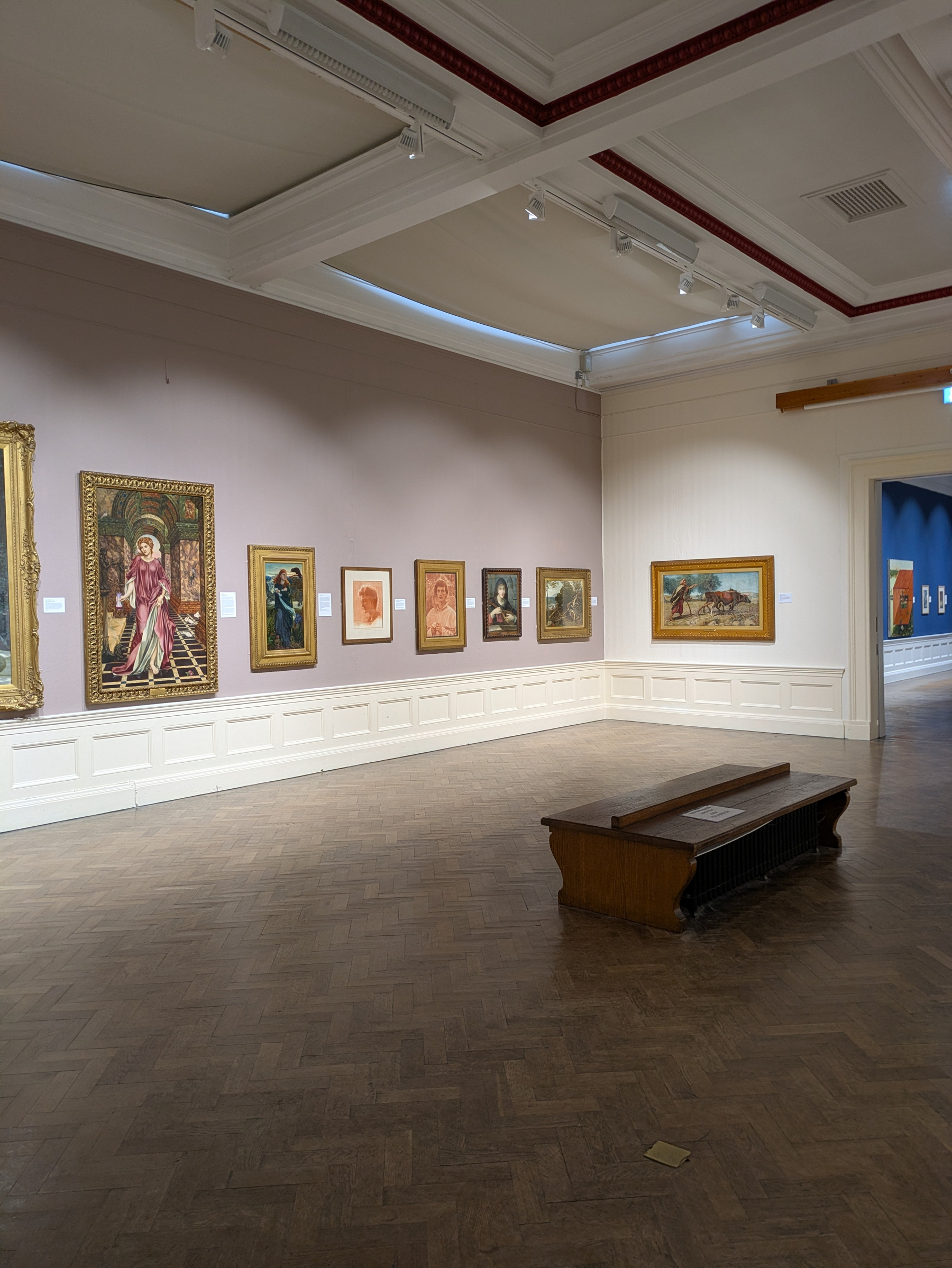 A gallery room displays various paintings on the walls with a wooden bench in the center.