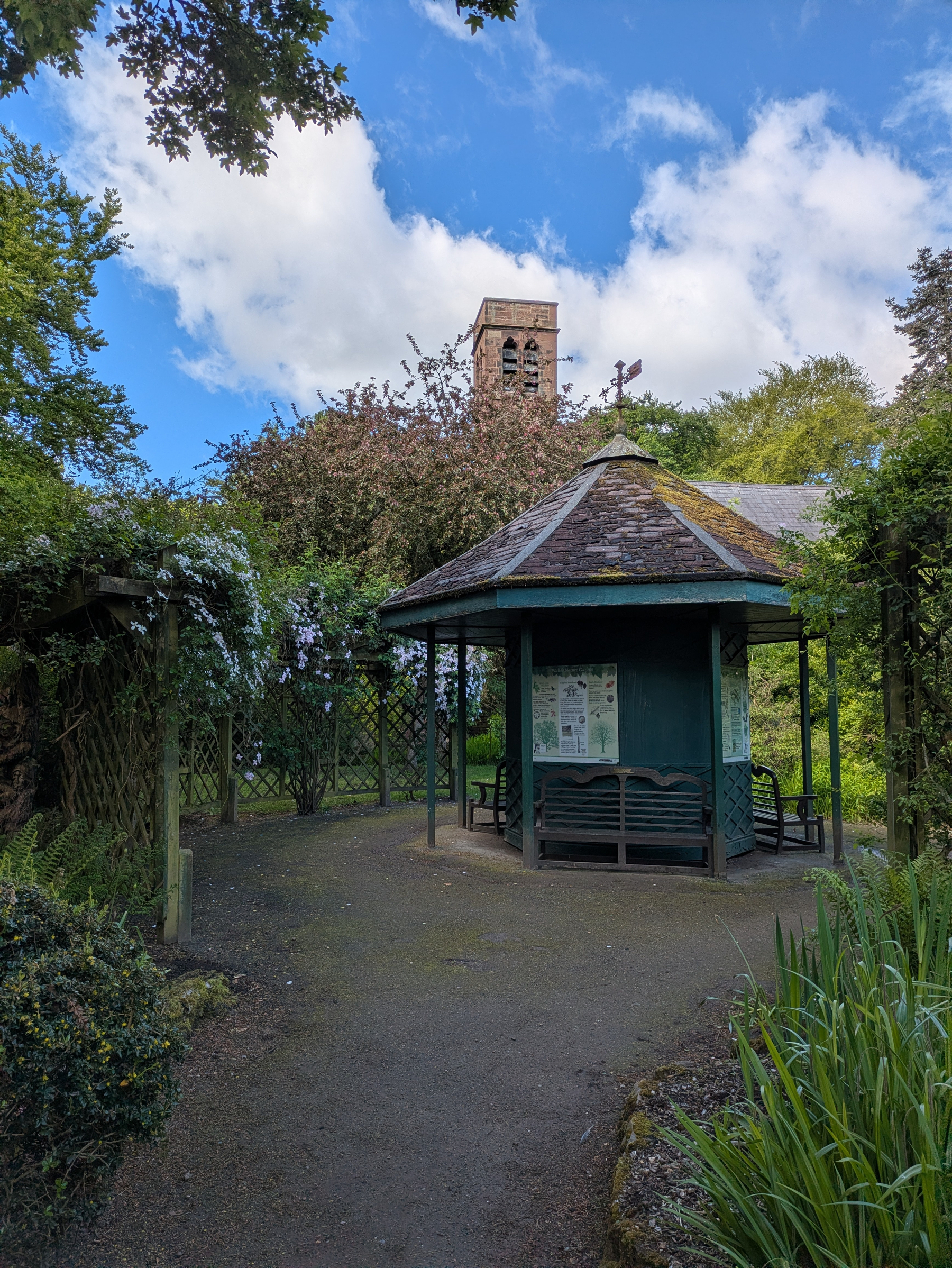 A small pavilion surrounded by greenery and trees, with a stone tower visible in the background under a partly cloudy sky.