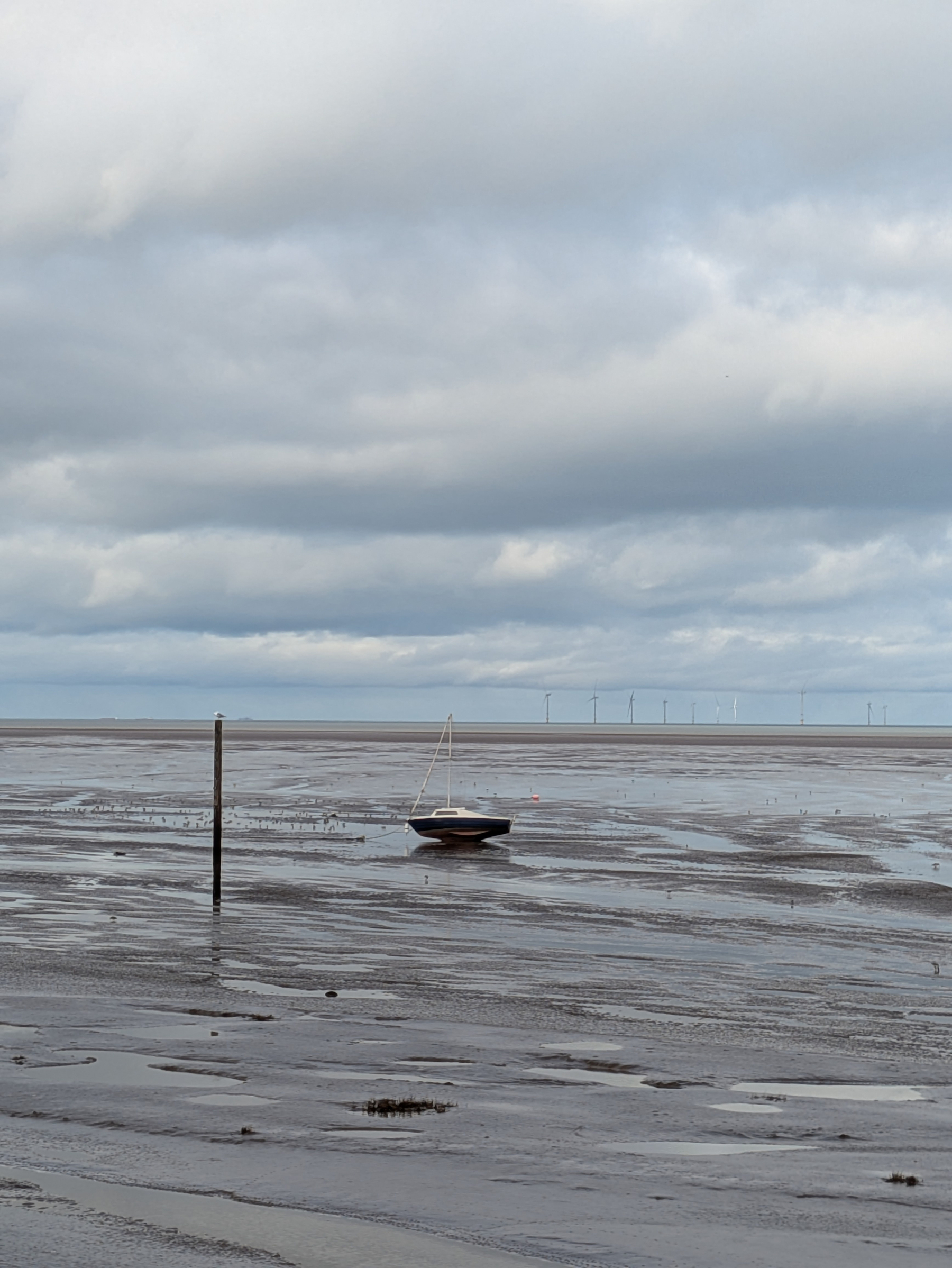 A small boat rests on a vast, muddy shoreline under a cloudy sky, with wind turbines visible in the distance.