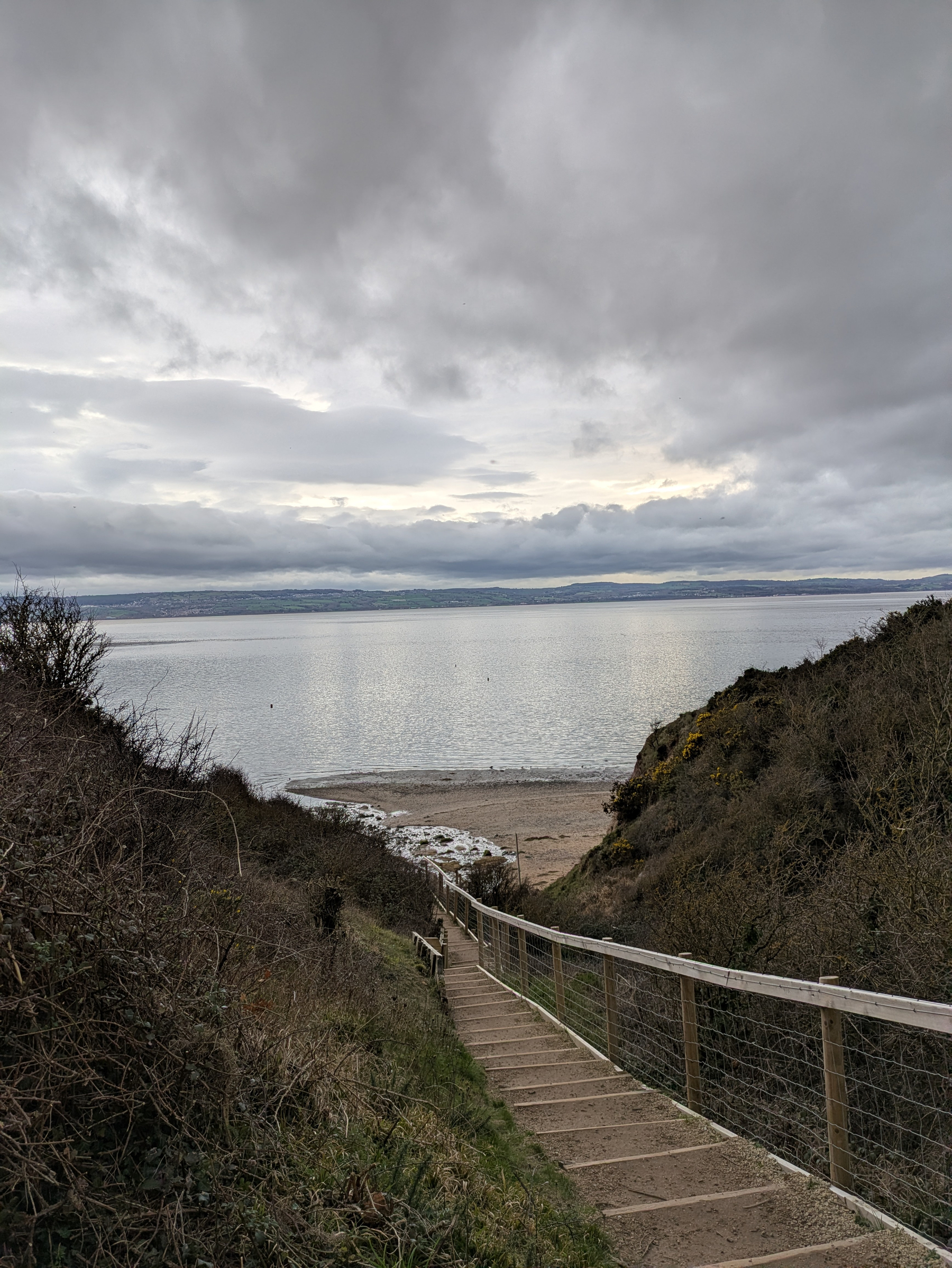 A wooden staircase leads down to a beach with a calm sea under a cloudy sky.