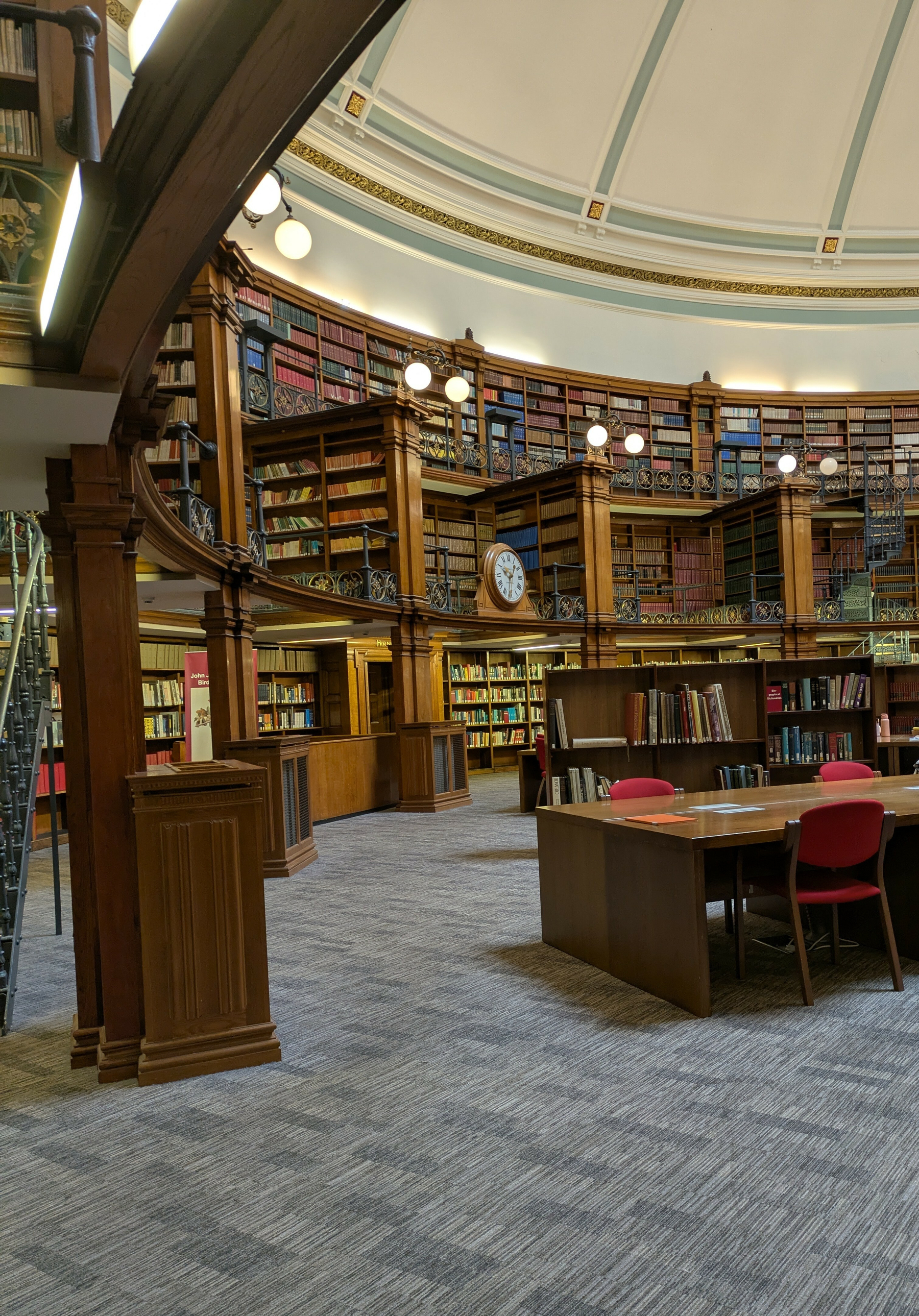 A large, ornate library room features curved bookshelves filled with books, a clock on the wall, and a table with chairs in the center.