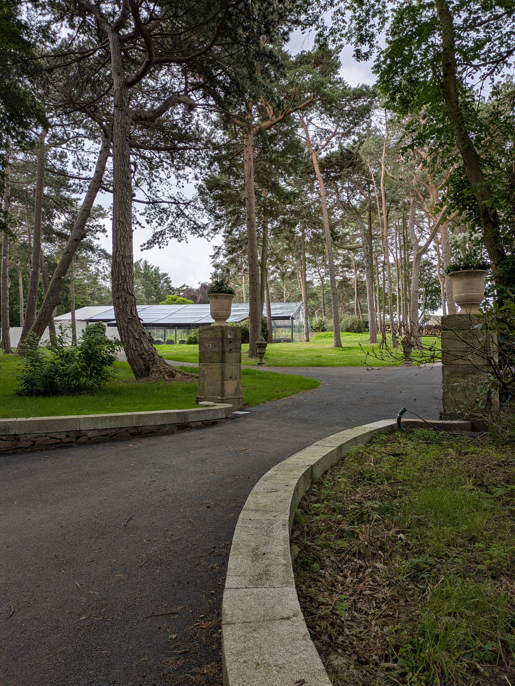 A tarmac pathway curves through a lush, green park surrounded by tall trees and stone pillars, with a function suite visible in the background.