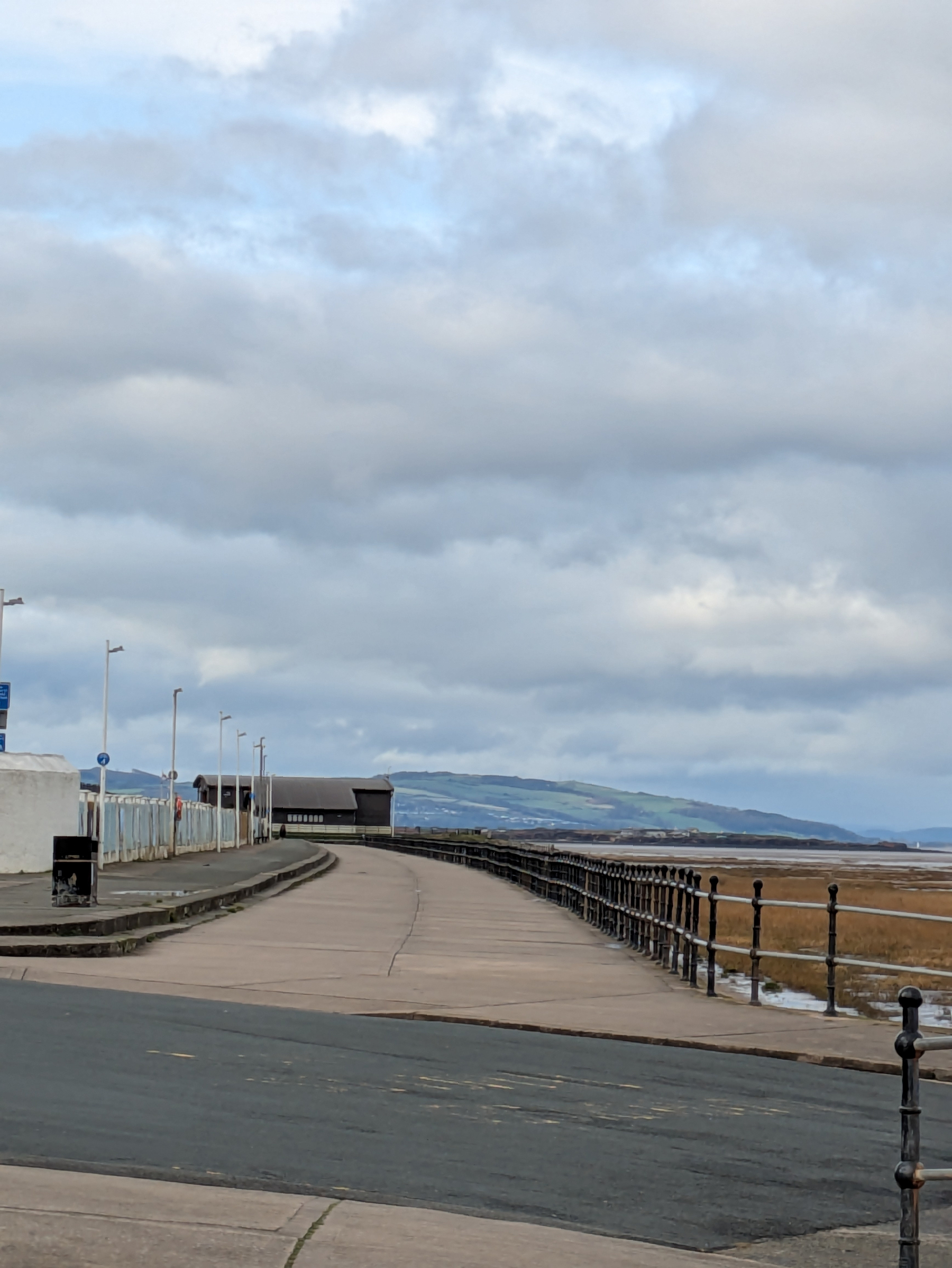 A coastal promenade with railings curves alongside a sandy beach under a cloudy sky.