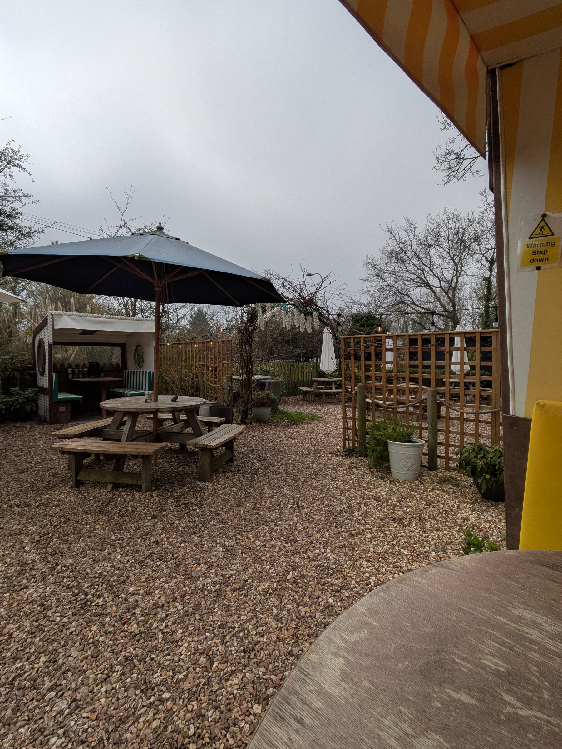 An outdoor seating area features wooden picnic tables, umbrellas, and a gravel ground surrounded by trees and fencing.