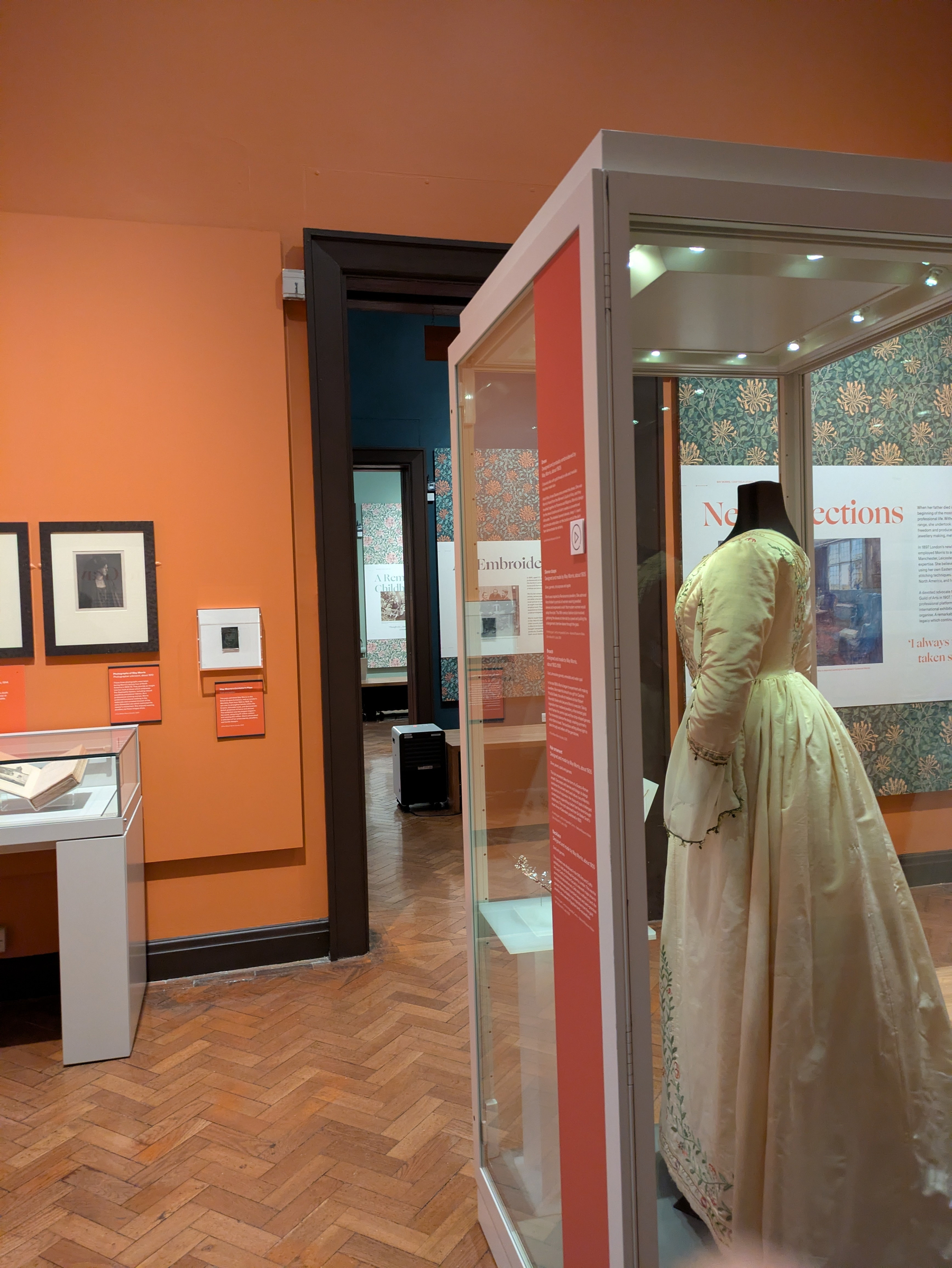 An exhibition room showcases vintage clothing on a mannequin within a glass case, alongside framed artworks and informational panels on the orange walls.