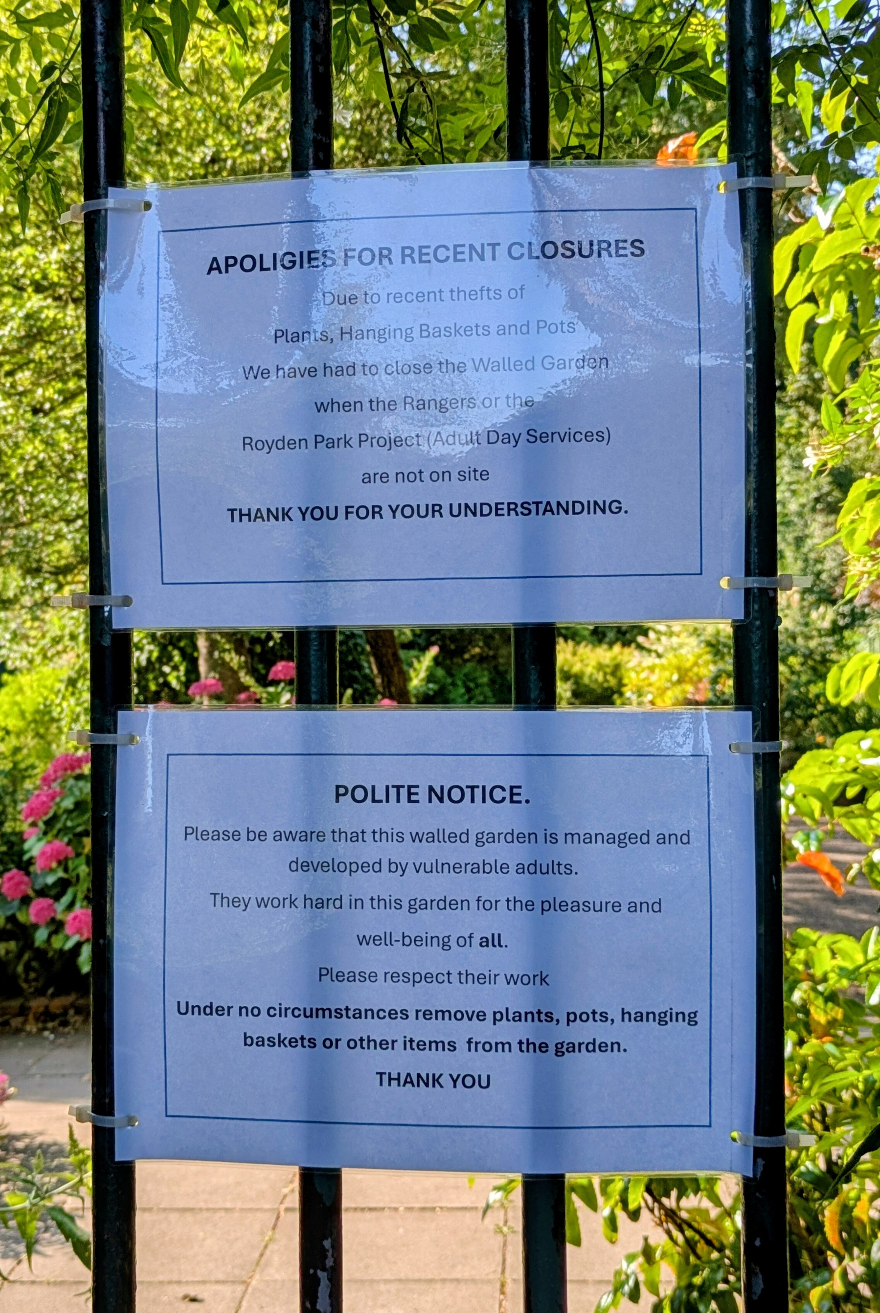 Two laminated signs on a black iron gate inform visitors about recent closures of a walled garden due to thefts and request respect for the work of vulnerable adults who maintain the garden.