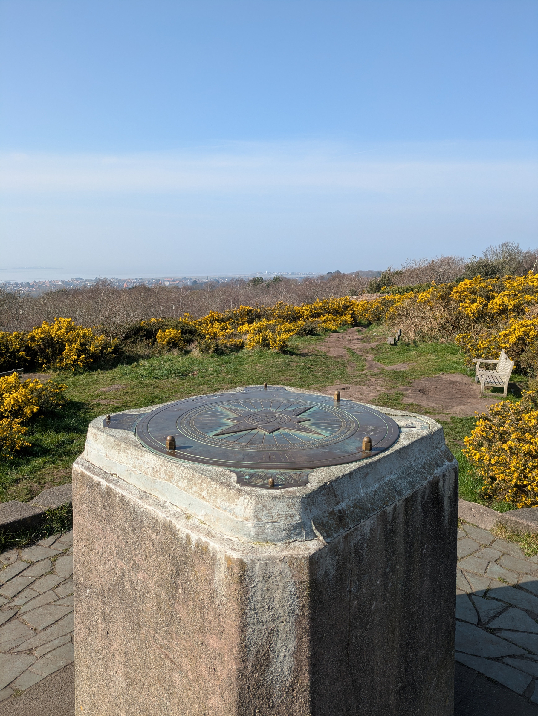 A large stone pedestal topped with a brass plaque featuring a compass design overlooks a scenic landscape with yellow flowering bushes and a bench in the distance under a clear blue sky.