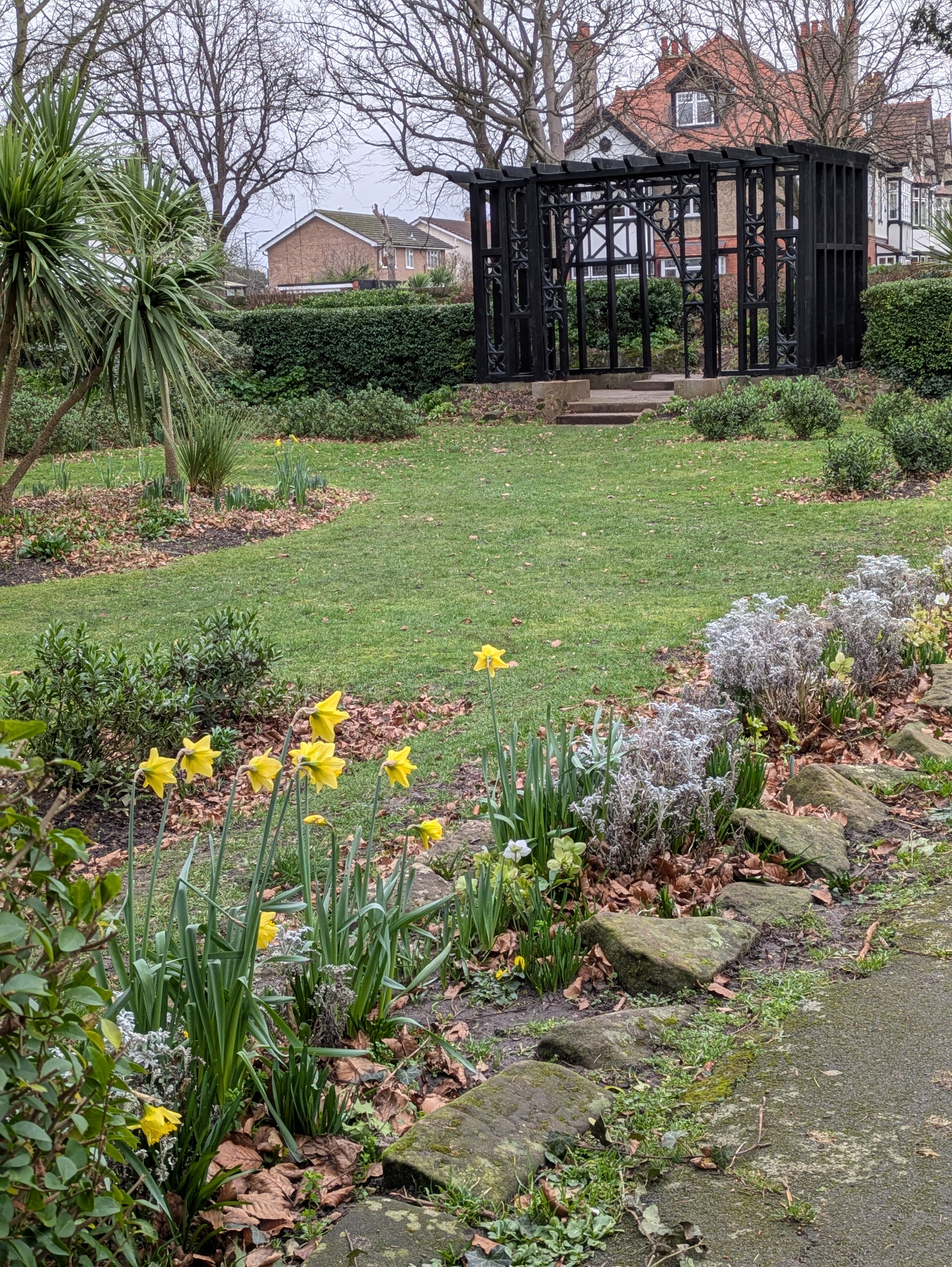 A garden scene features daffodils, a manicured lawn, and a black wooden structure in the background, bordered by trees and shrubs.