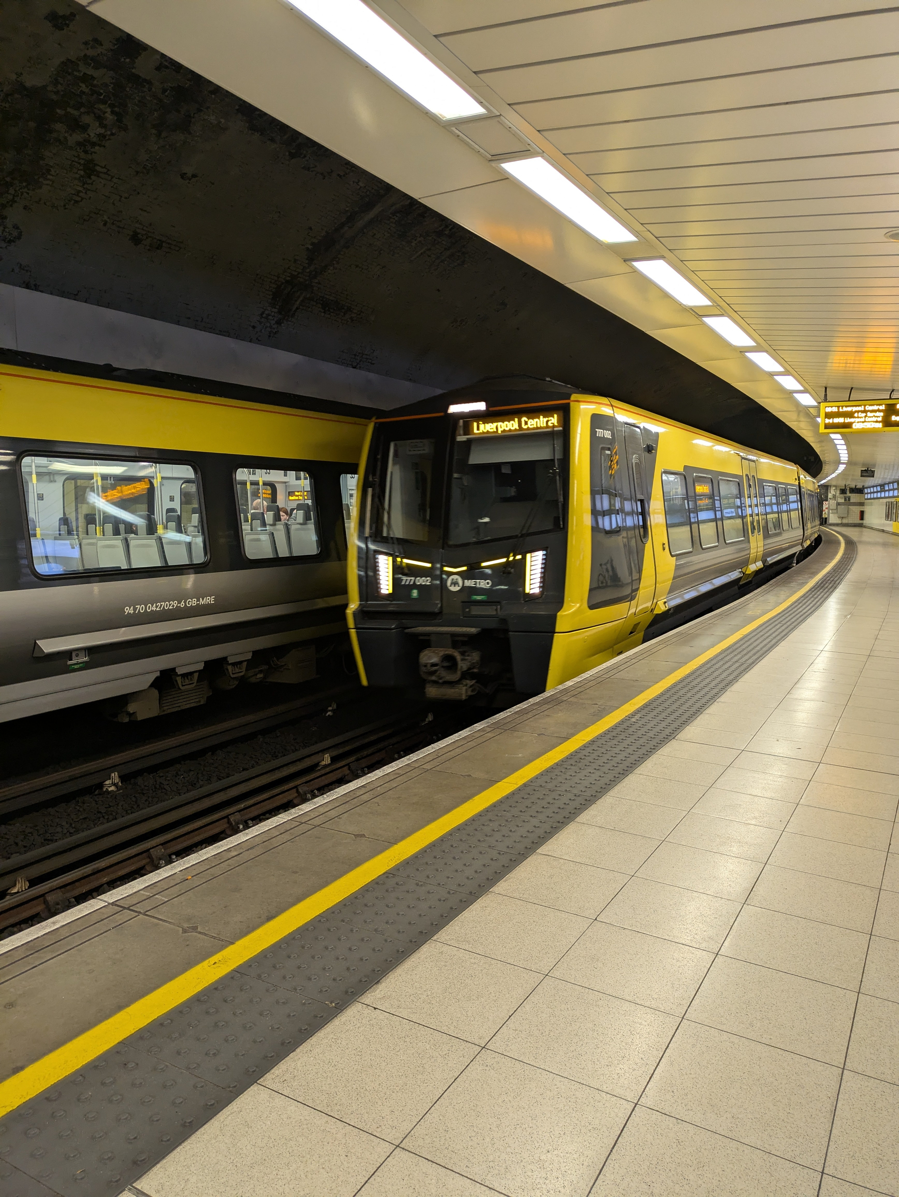 A modern yellow and gray train arrives at an underground station platform with bright overhead lighting.