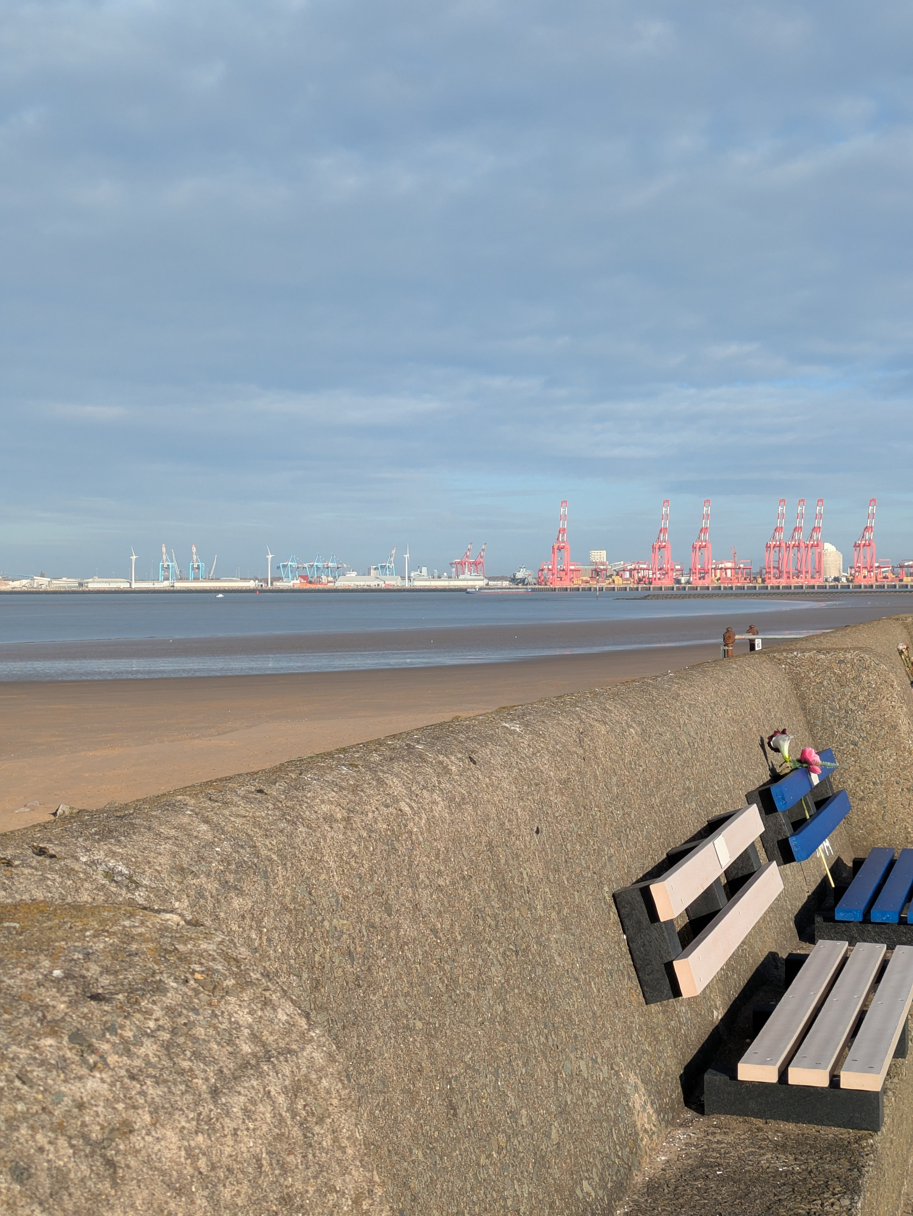 A coastal scene features a concrete barrier with colorful wooden benches overlooking a sandy beach and industrial cranes in the background.