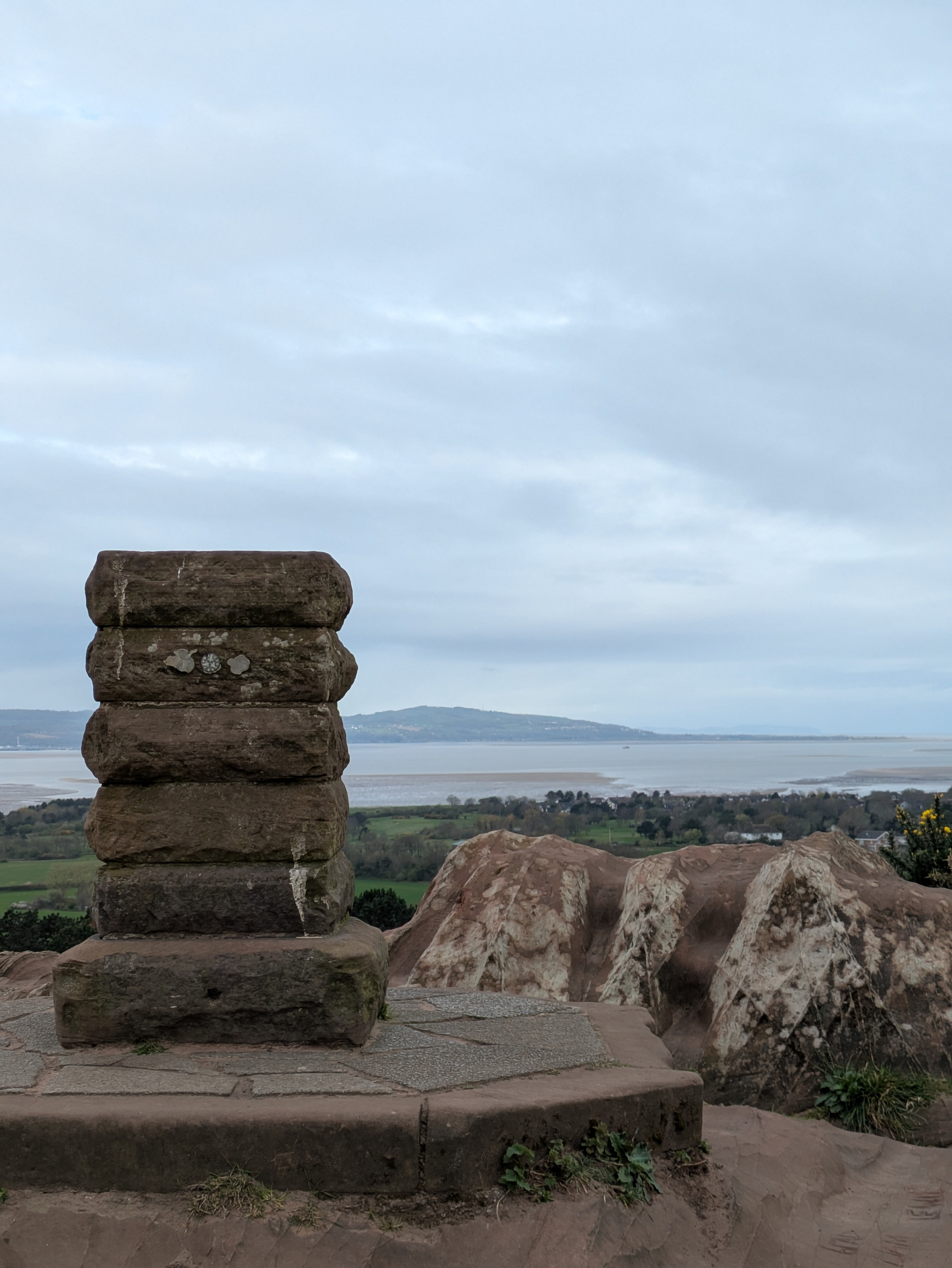 A stone monument stands on a rocky hill overlooking a coastal landscape with water and distant landforms under a cloudy sky.