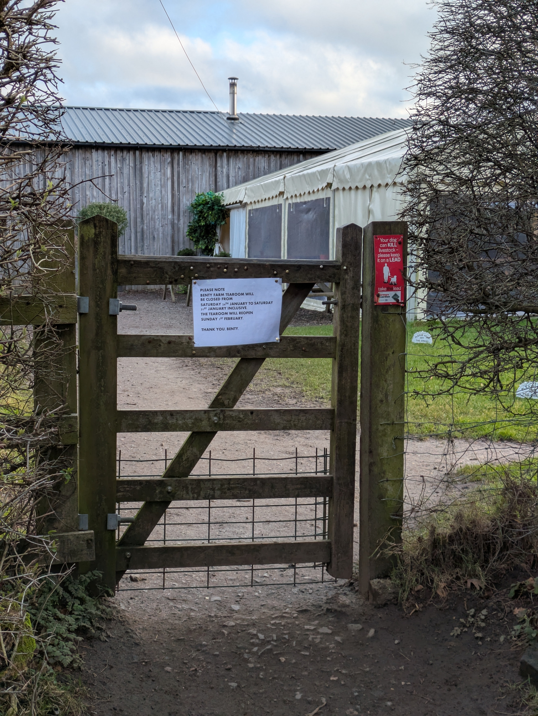 A wooden gate leads to a path flanked by bushes, with a sign attached and a building and white tent visible in the background.