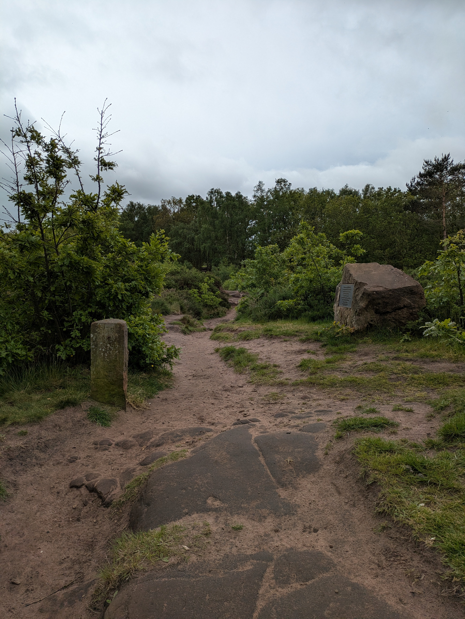 Dirt path leading past boundary marker and memorial stone with treeline in distance.