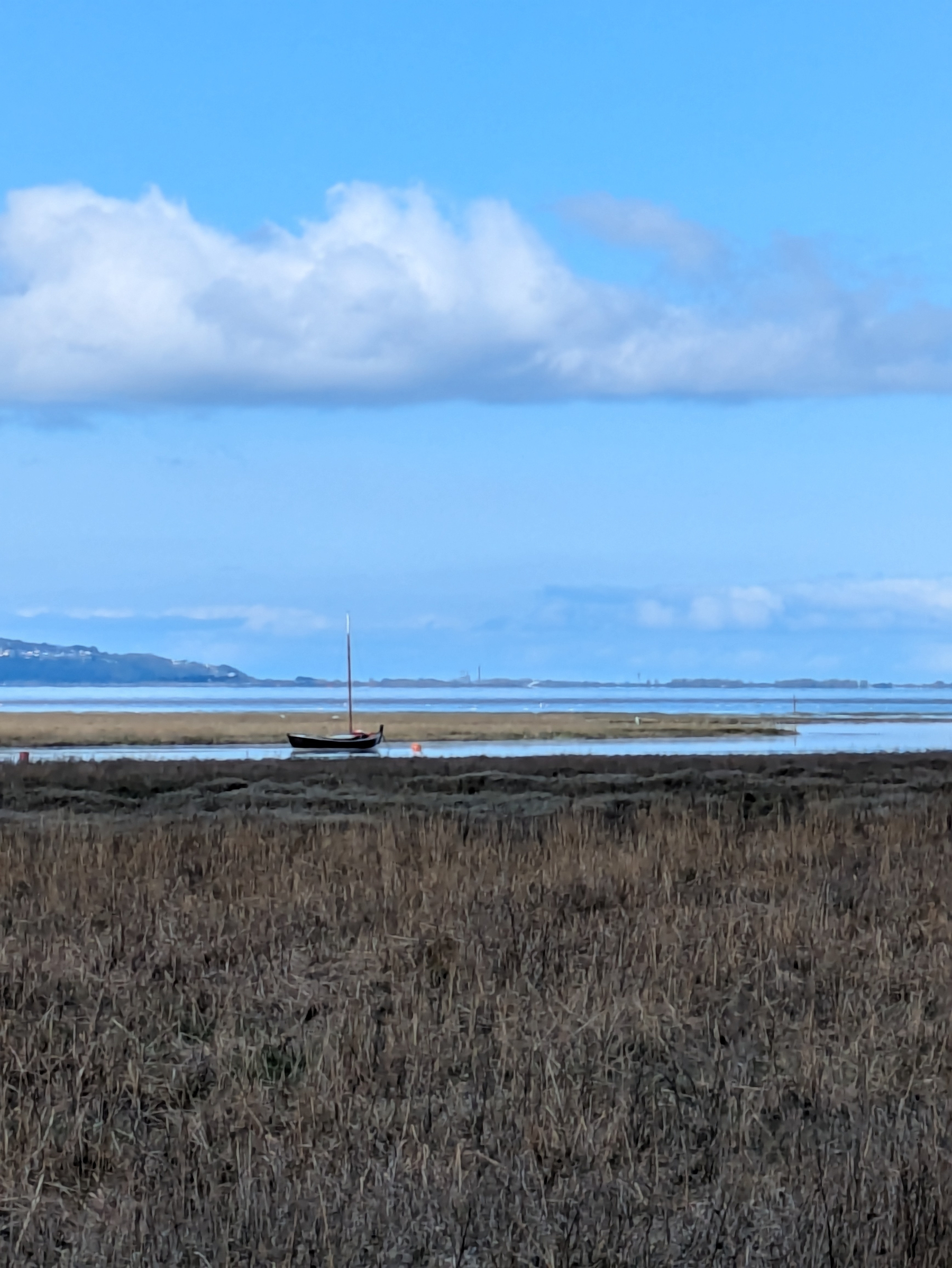 A sailboat rests on calm waters under a blue sky with scattered clouds, surrounded by a tranquil landscape.