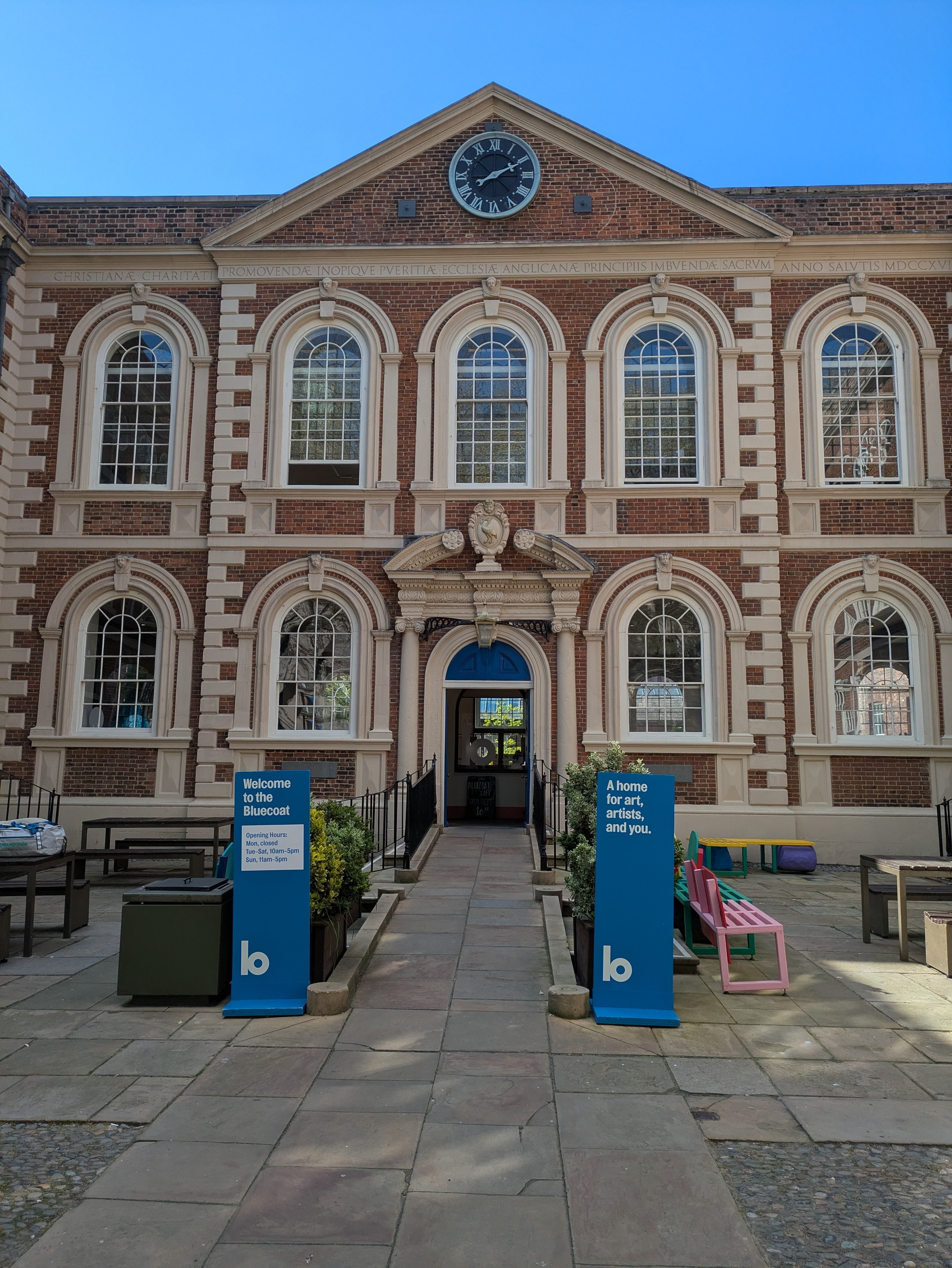 A historic brick building featuring arched windows and a clock is fronted by informational blue signs and outdoor seating in a courtyard.