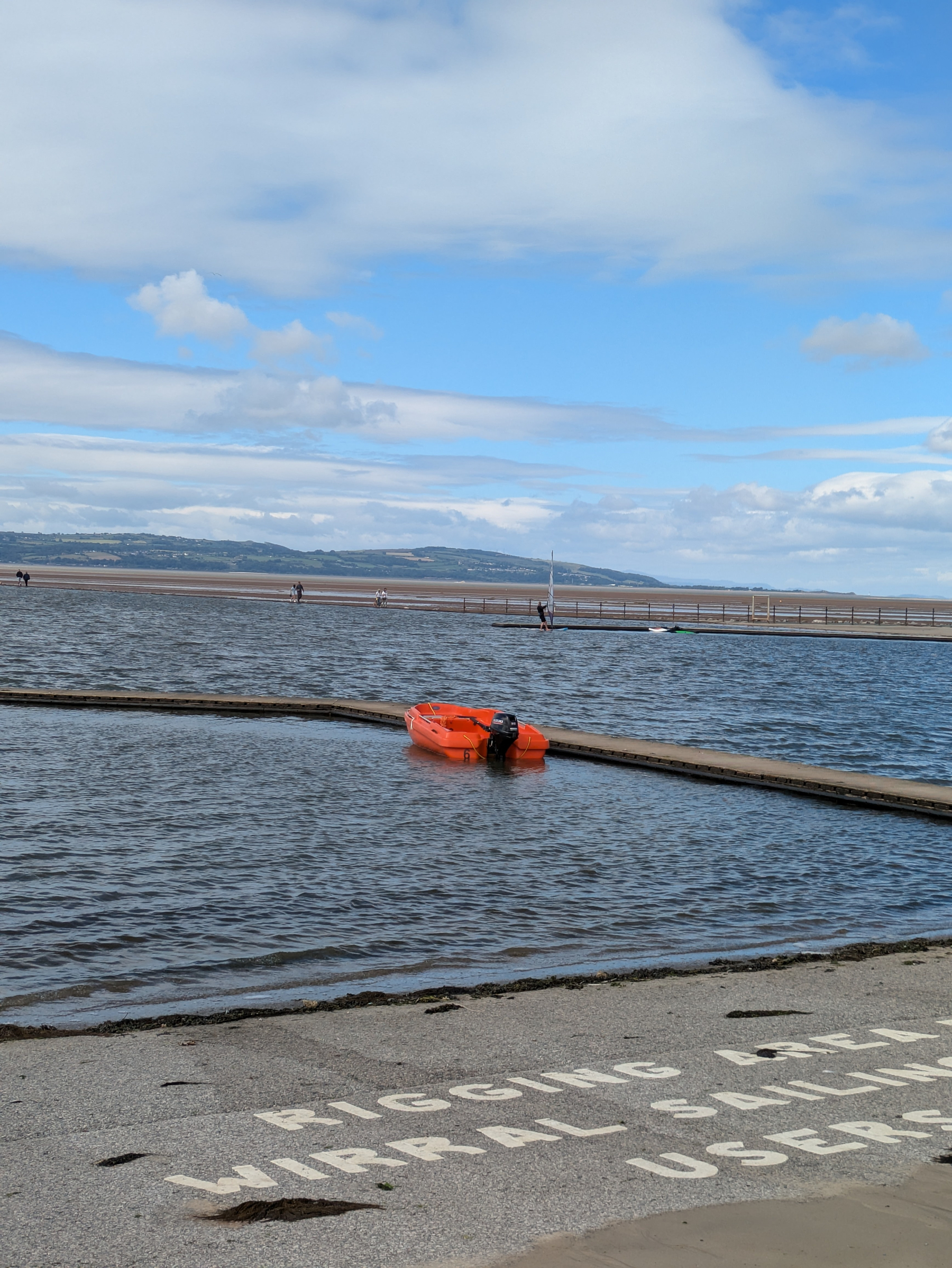 A solitary orange boat floats in a calm, shallow body of water under a partly cloudy sky, with distant land visible across the horizon.