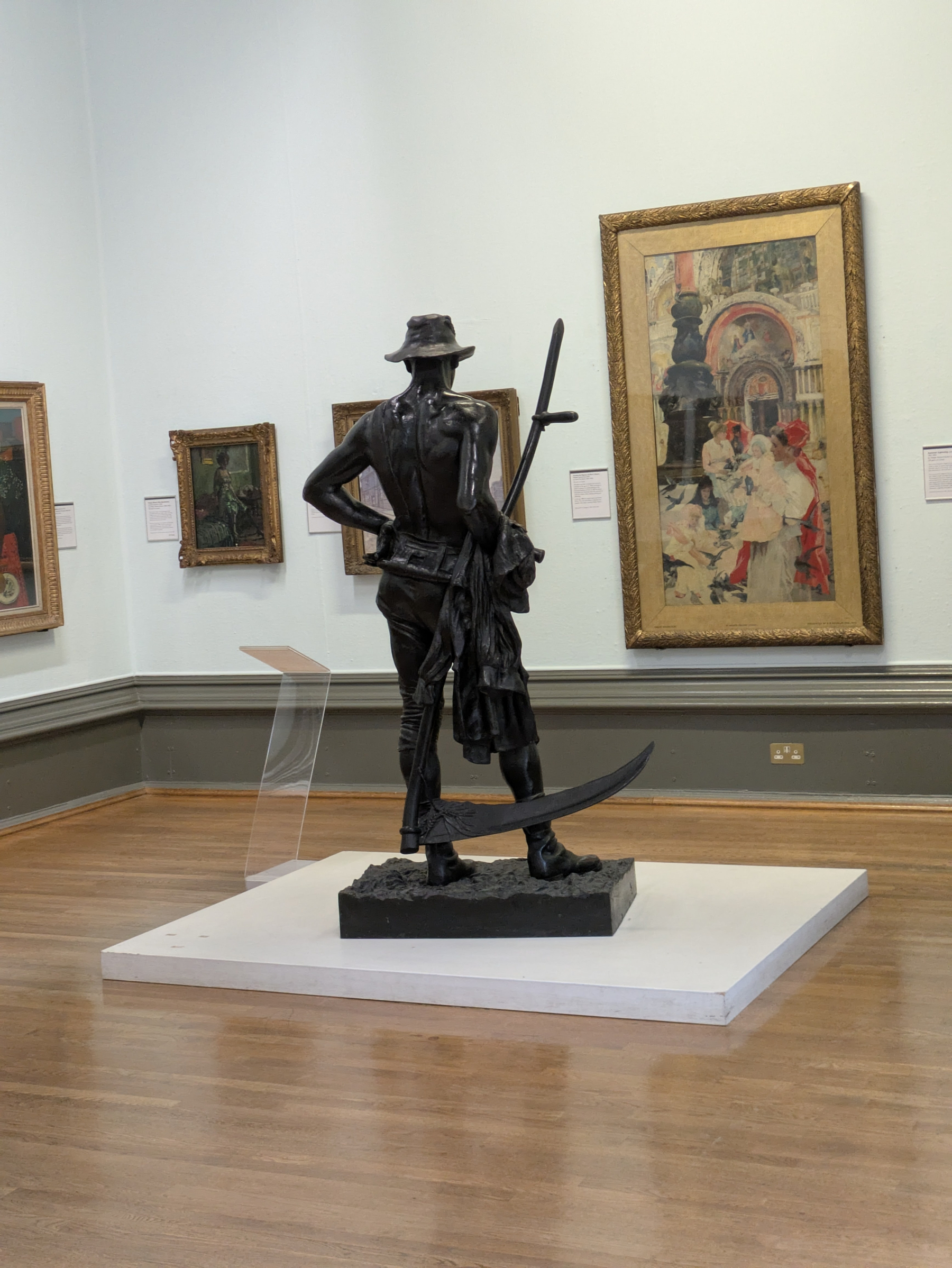 A bronze statue of a farmworker with a broad-brimmed hat stands on a pedestal in an art gallery, surrounded by framed paintings on the walls.