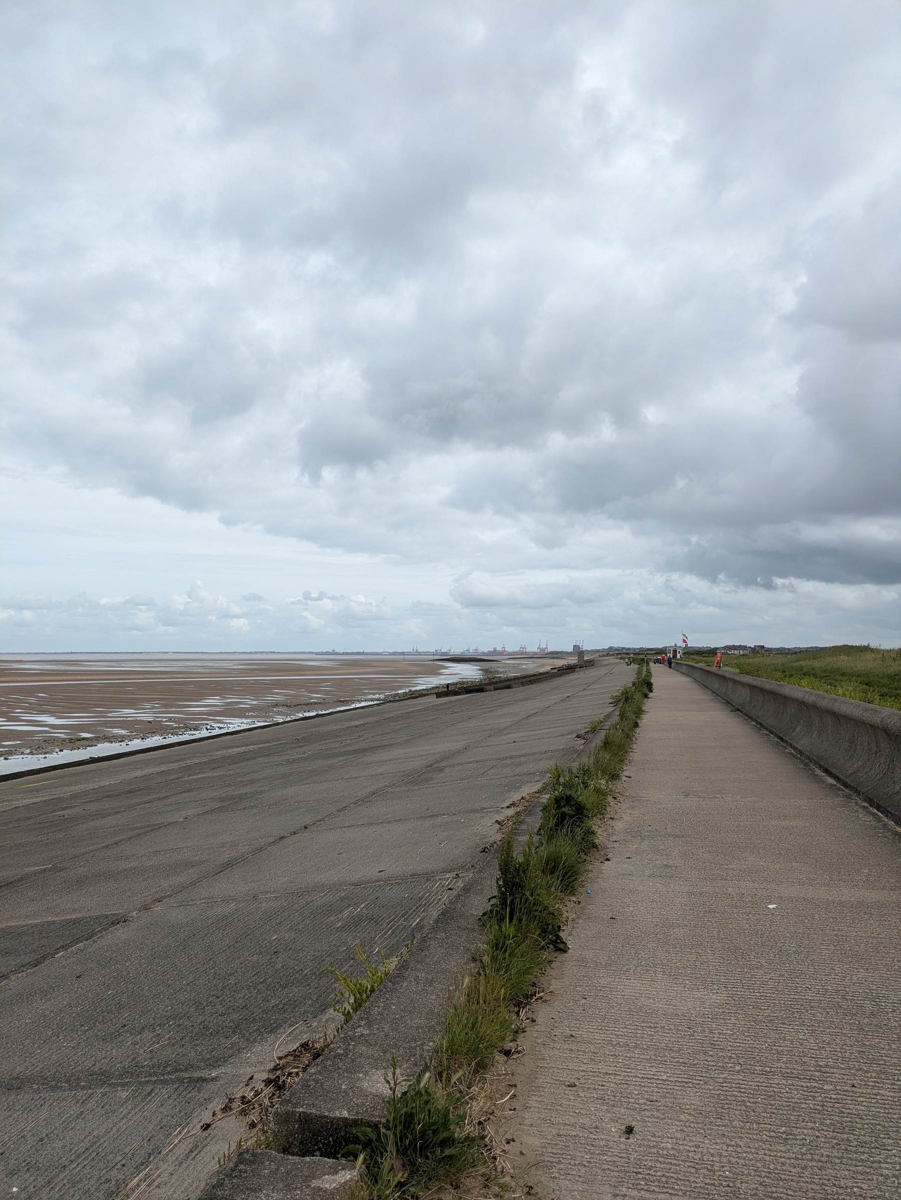 A paved pathway runs parallel to a sandy shoreline under a cloudy sky, with sparse greenery along the edge.