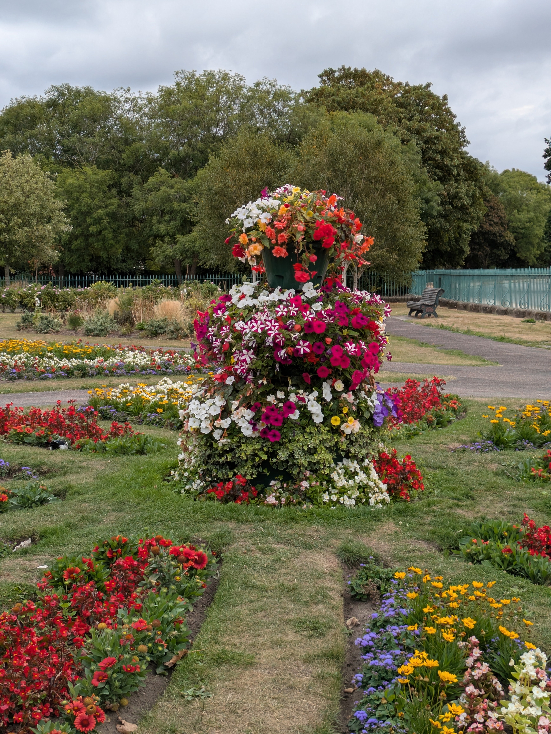 A vibrant floral arrangement shaped like a tiered tower stands in the center of a garden surrounded by colorful flower beds and greenery.