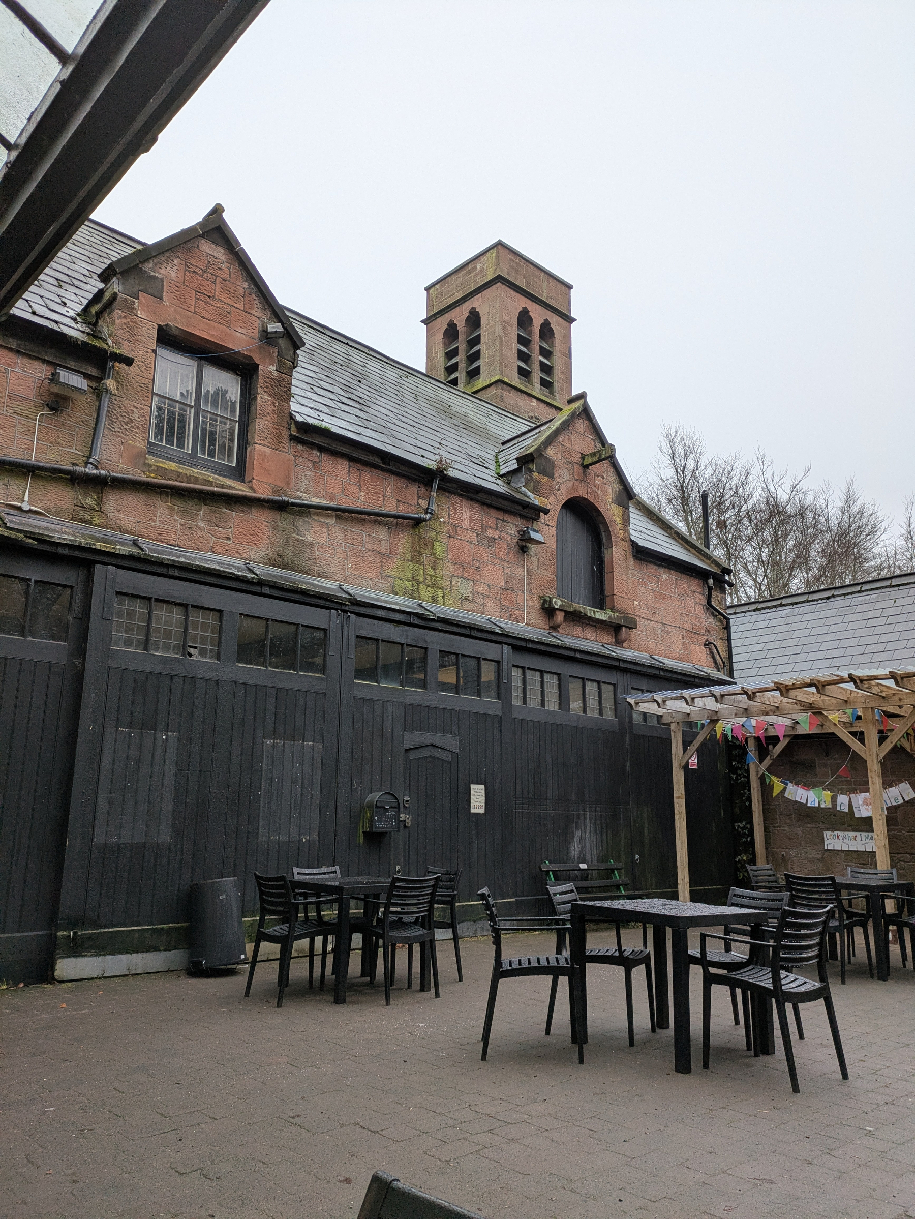 An outdoor patio with black tables and chairs is set against a rustic brick building with a small bell tower and slate roof.