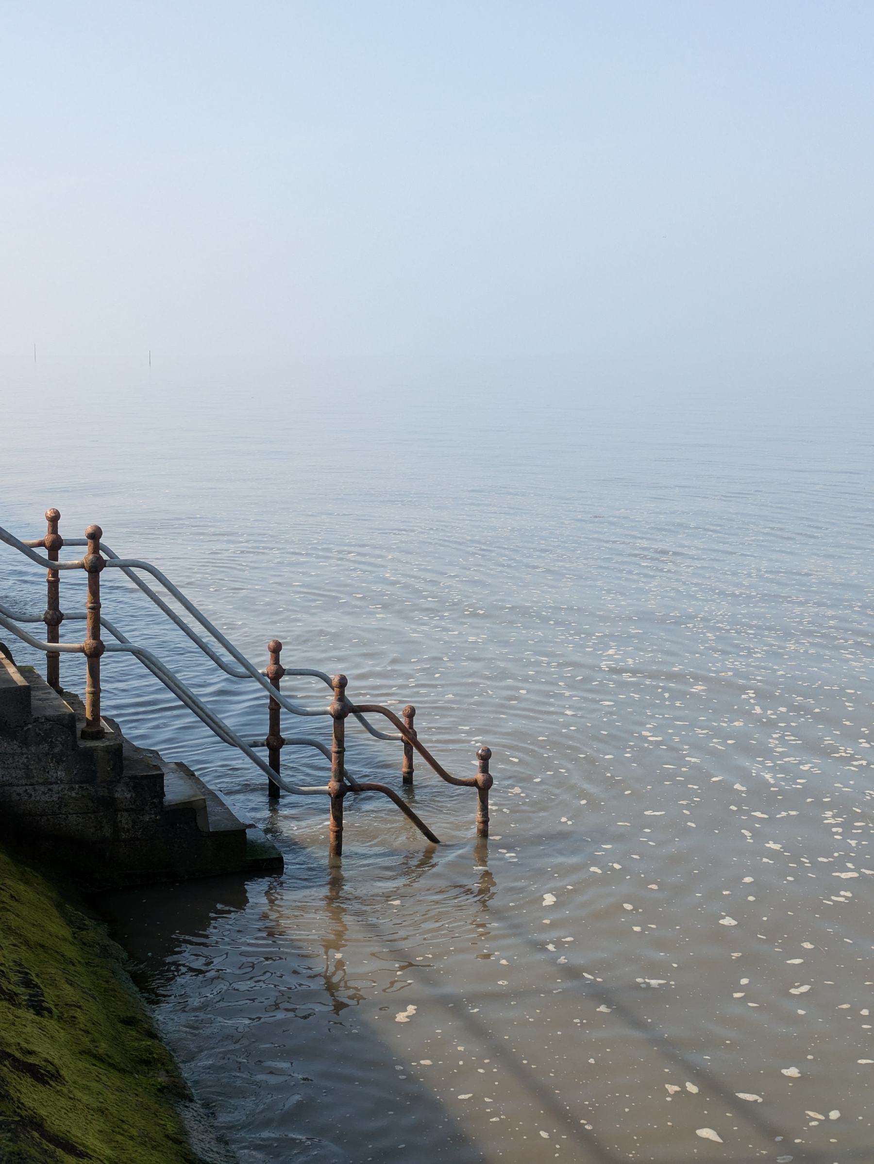 A metal railing and steps descend into calm, foggy waters under a hazy sky.