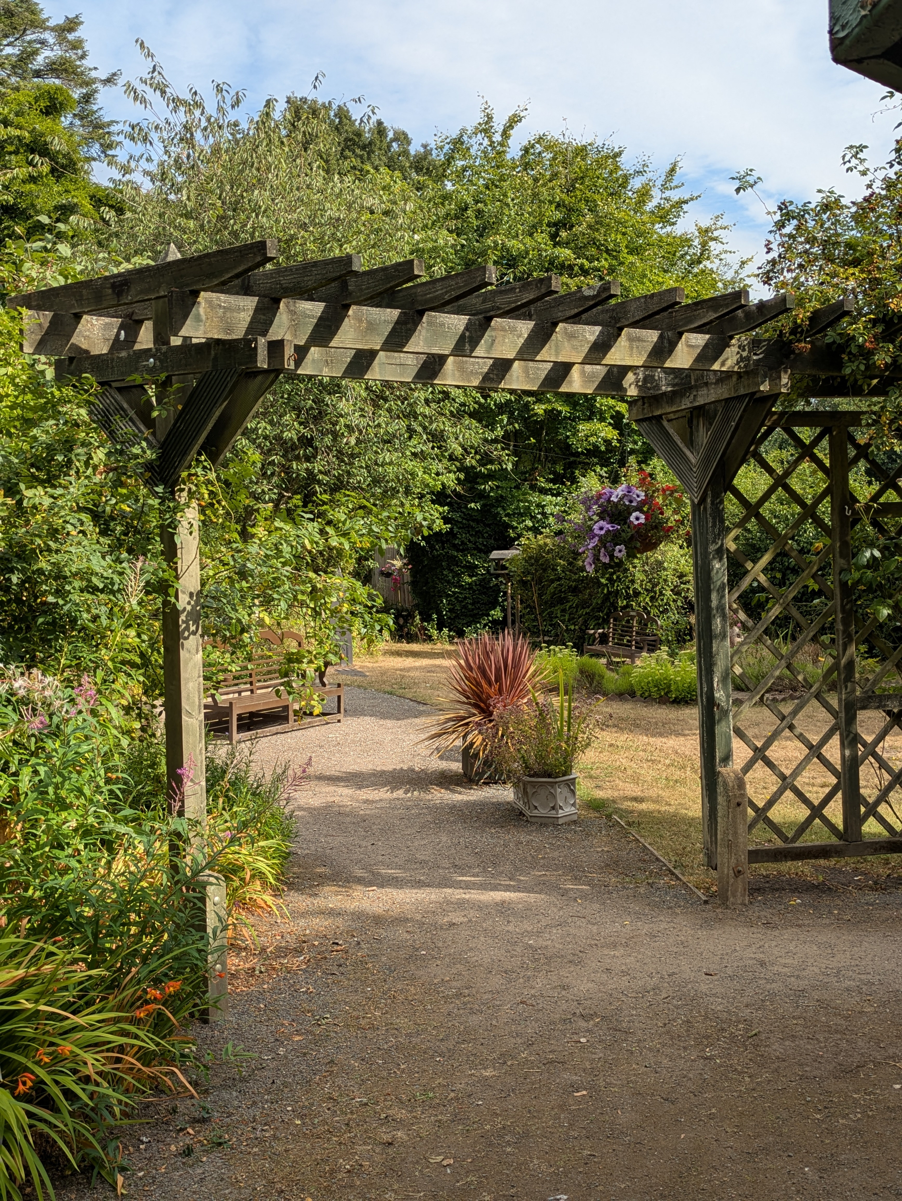 A wooden garden arbor stands over a gravel pathway surrounded by lush greenery and colorful plants.