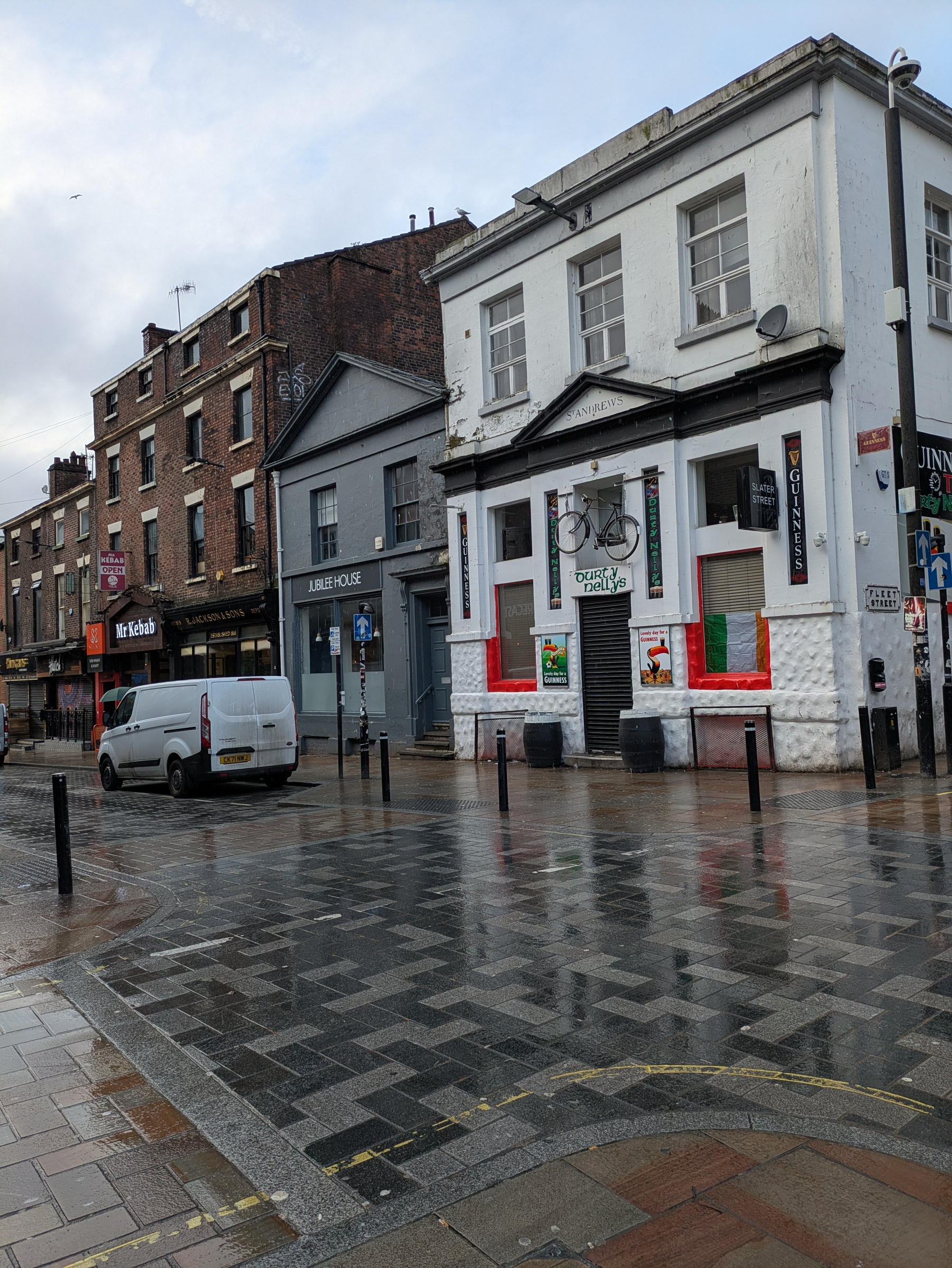 A street scene features a white building with a bicycle mounted above the door, surrounded by brick buildings and wet pavement.