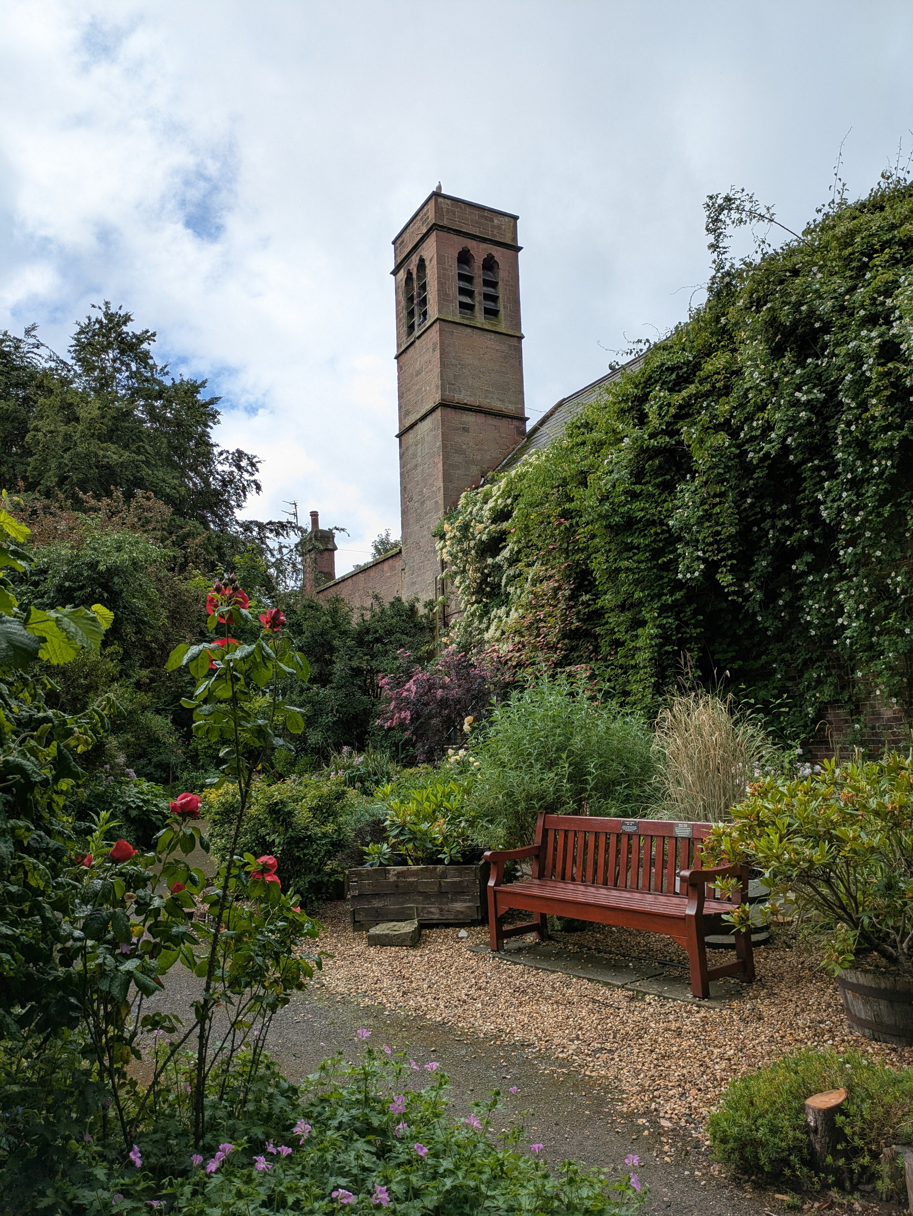 A peaceful garden scene features a wooden bench, vibrant flowers, lush greenery, and a tall brick tower in the background.