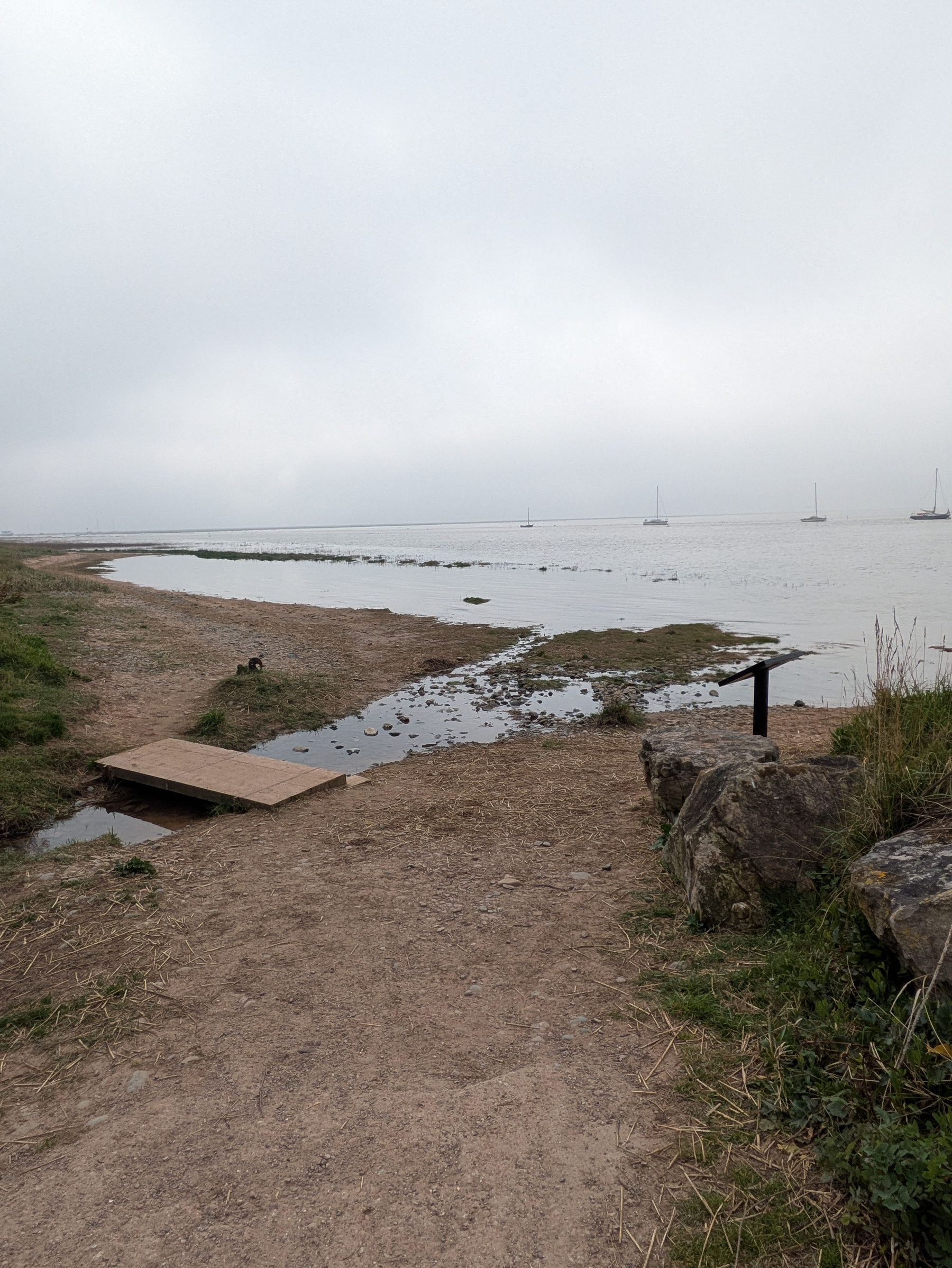 A sandy coastal path leads to a misty shoreline with sailboats in the distance.