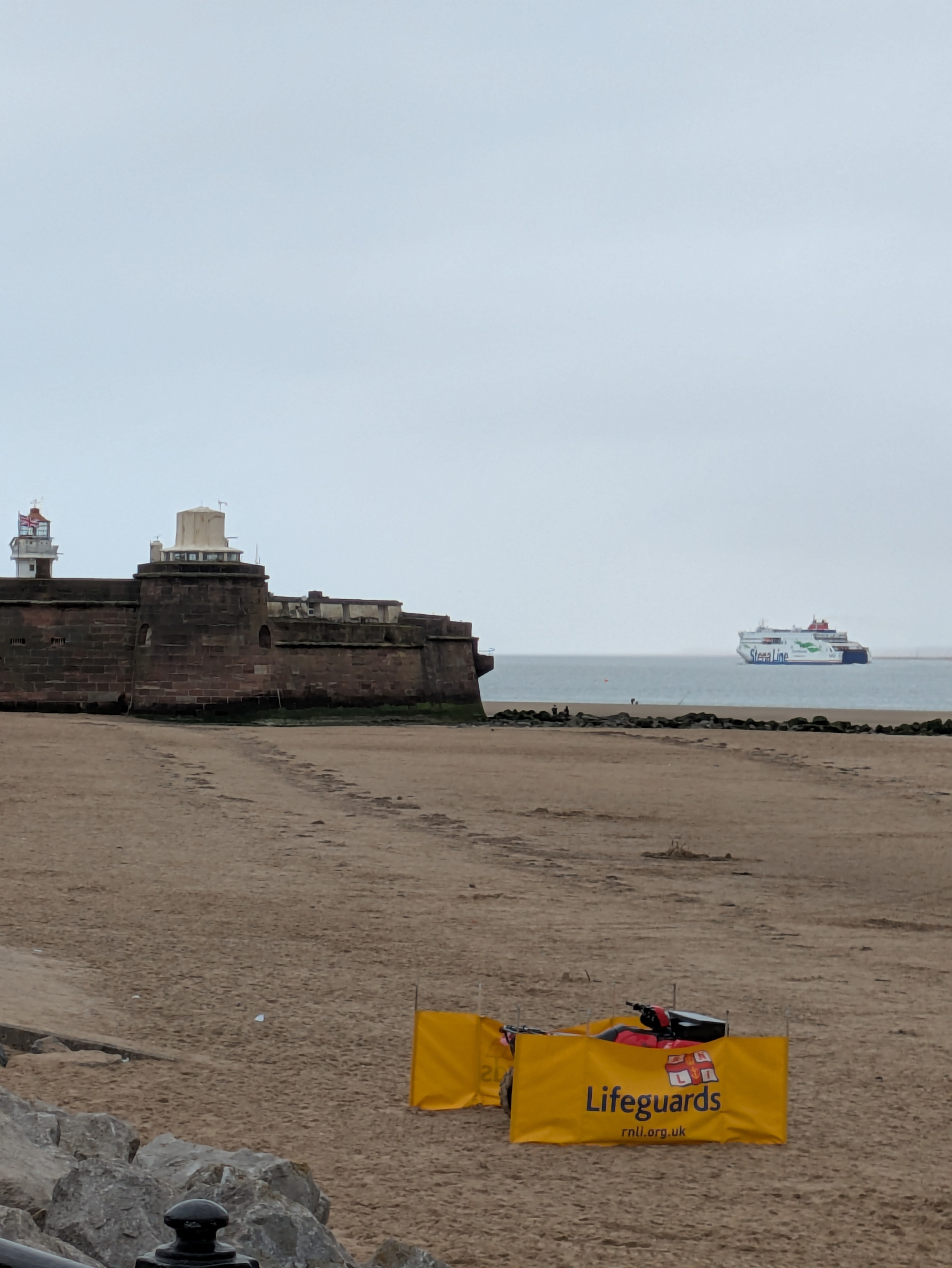 A sandy beach with a Lifeguards sign and a distant ferry near a stone fortification and lighthouse by the sea.