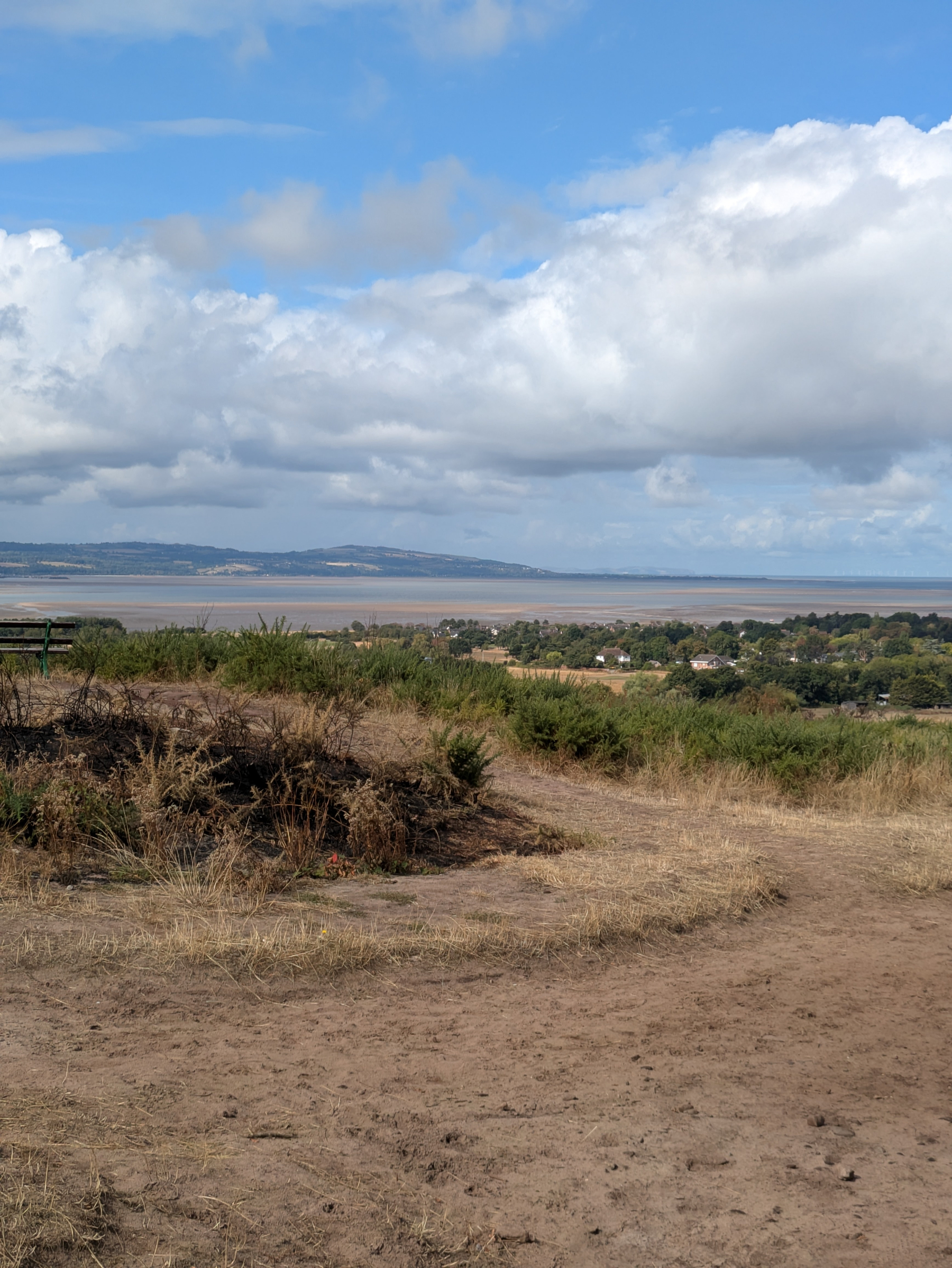 A scenic view overlooking a grassy, slightly barren hillside with a bench on the left, leading to a small town and a body of water under a partly cloudy sky.