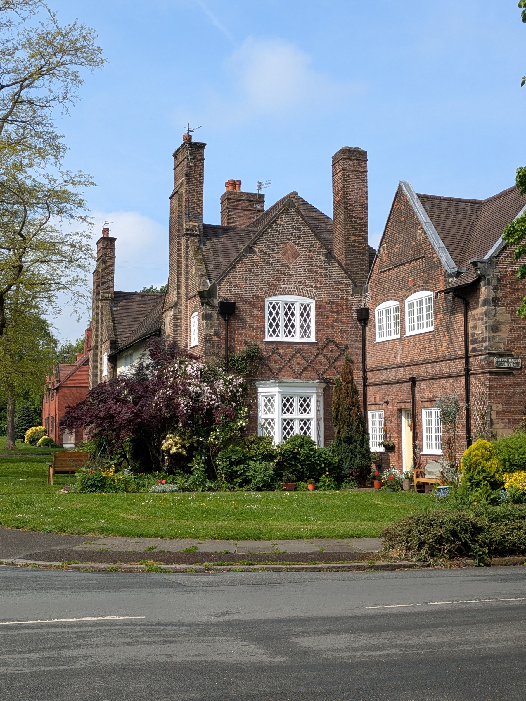 A charming brick house with tall chimneys and a lush garden sits near the roadside.