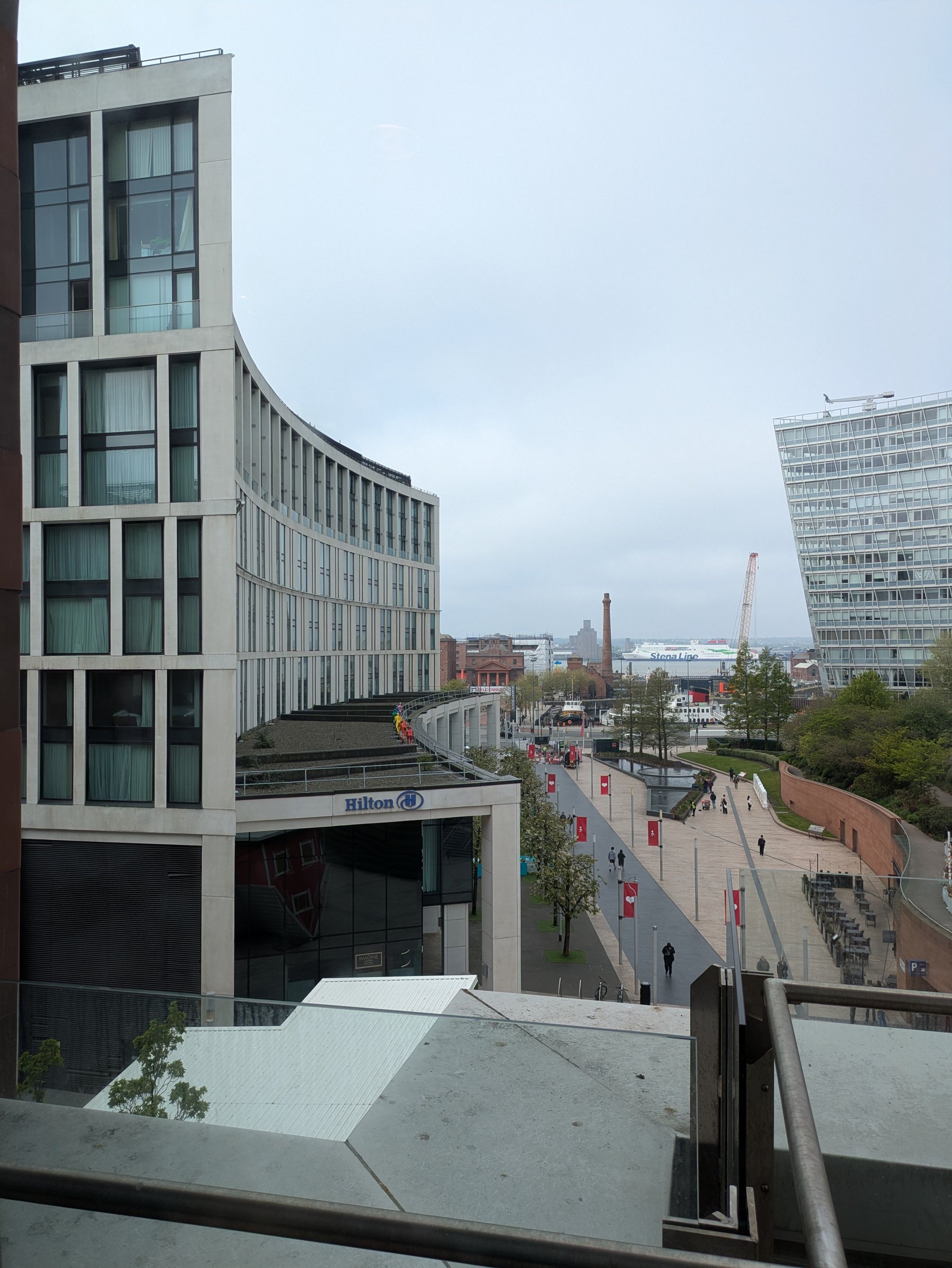 The view features a modern urban area with tall buildings, a pathway lined with flags, and a cloudy sky above.