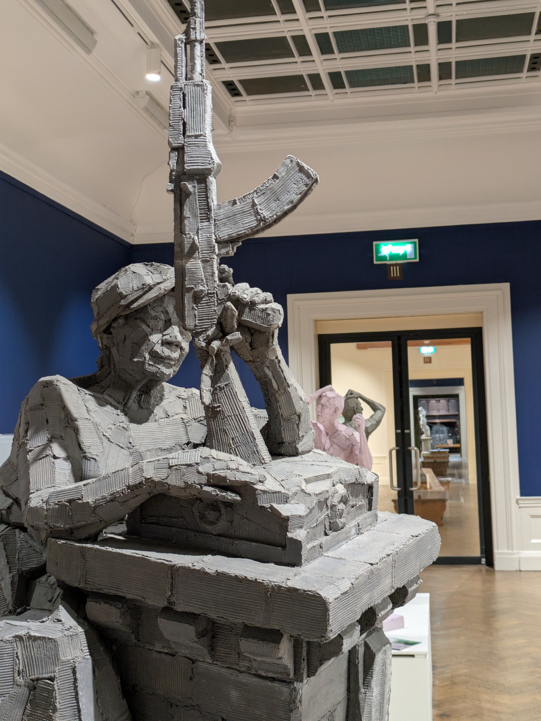 A detailed sculpture in a gallery depicts a figure holding a large rifle, surrounded by a setting with a blue wall and a ceiling grid.