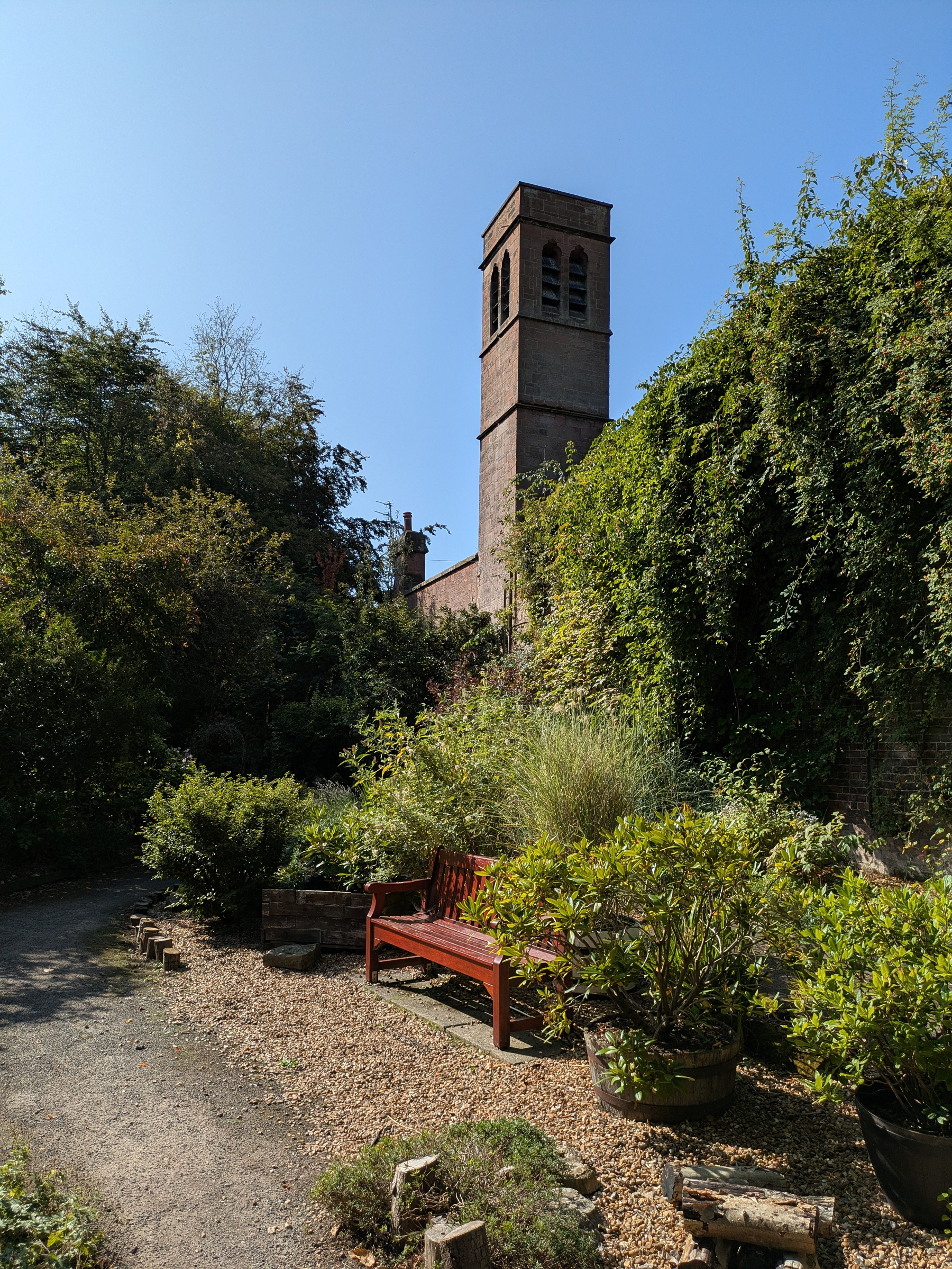 A tall brick tower rises above a lush garden area with a wooden bench and various green plants under a clear blue sky.
