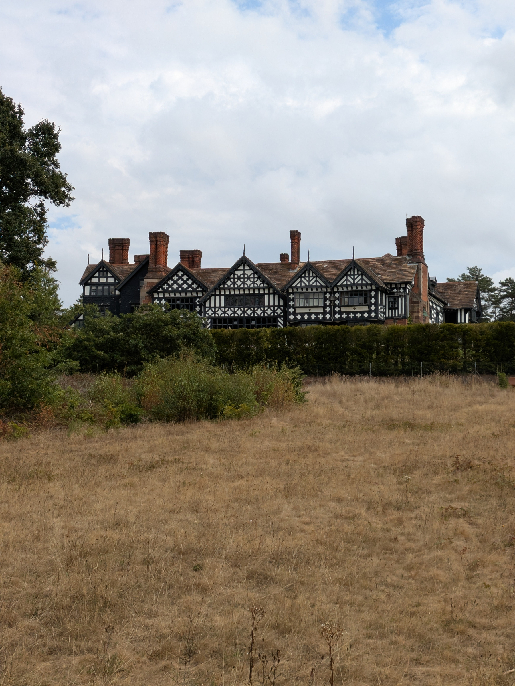 A large Tudor-style house with distinctive black and white timber framing is set behind a hedge with dry grass in the foreground.