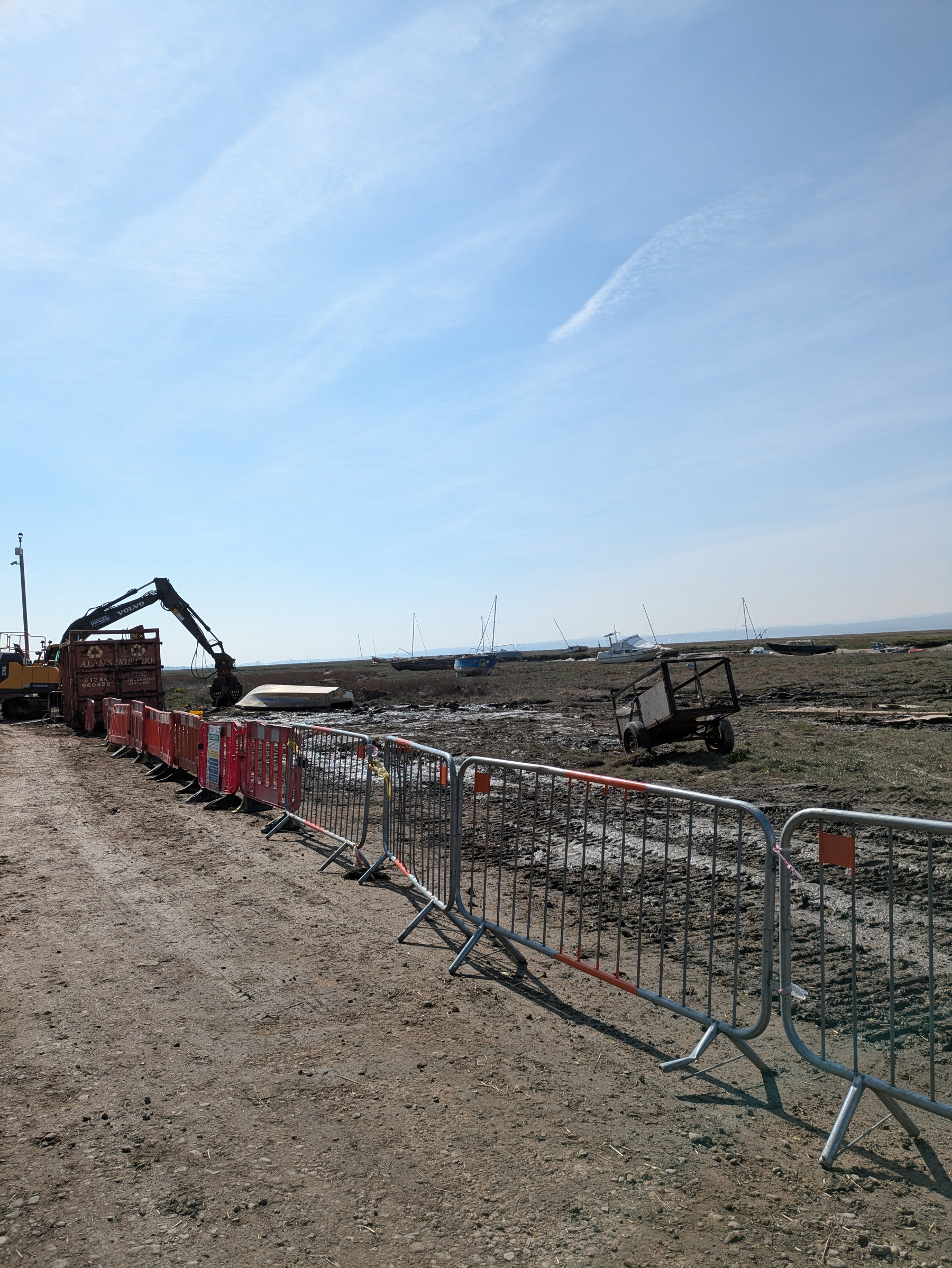 A construction site with a digger and various construction barriers is set against a backdrop of boats on a muddy shoreline.