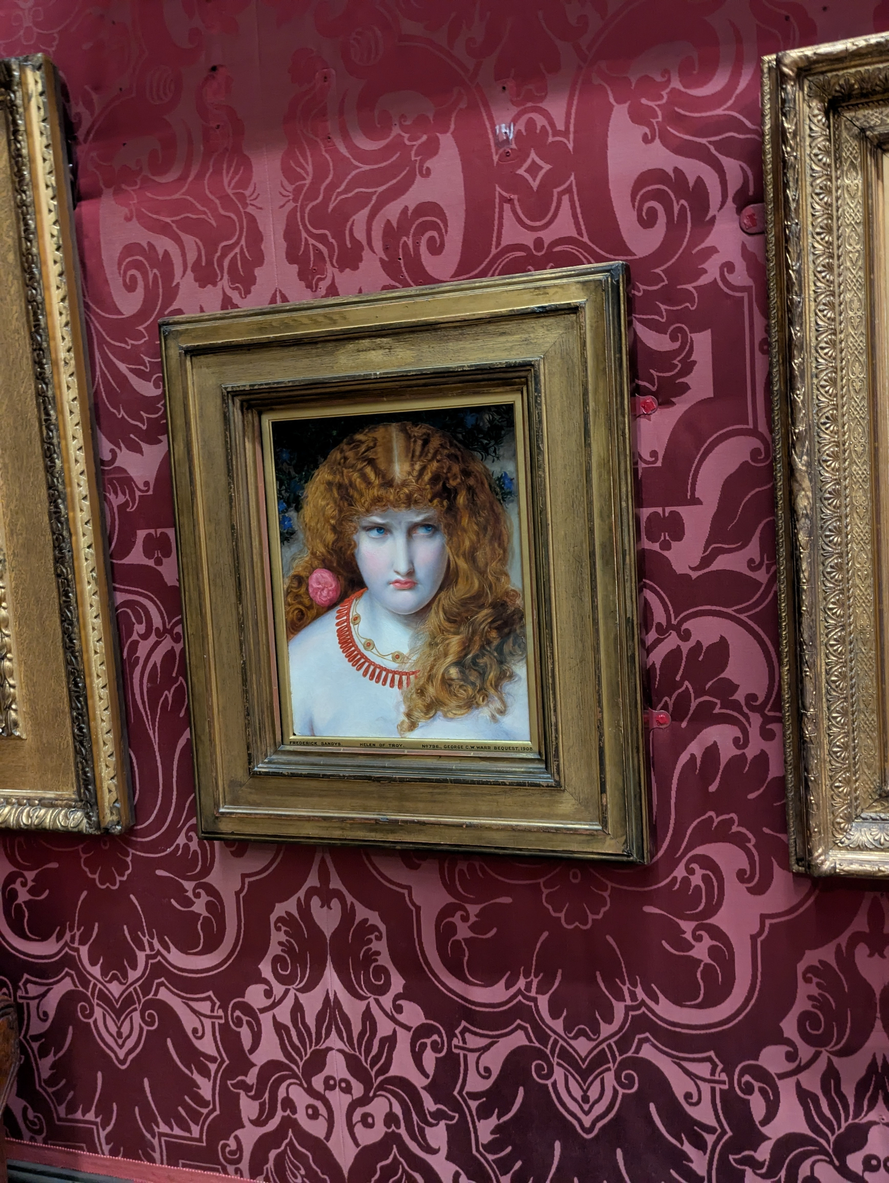 A framed painting of a long-haired individual with red jewelry is displayed on a wall with ornate red wallpaper.