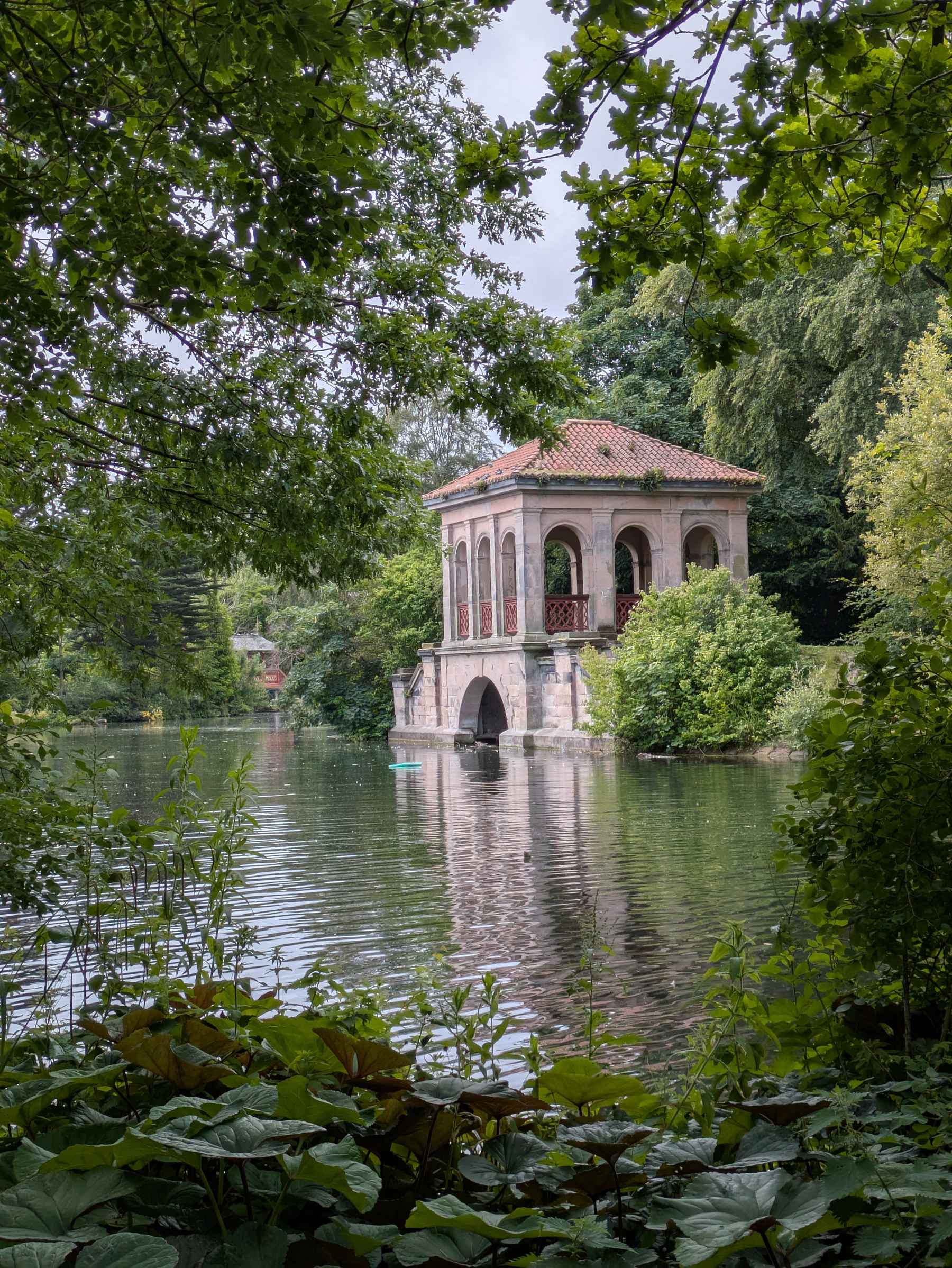 A small, picturesque building with arches and a tiled roof stands beside a serene lake, surrounded by lush greenery.