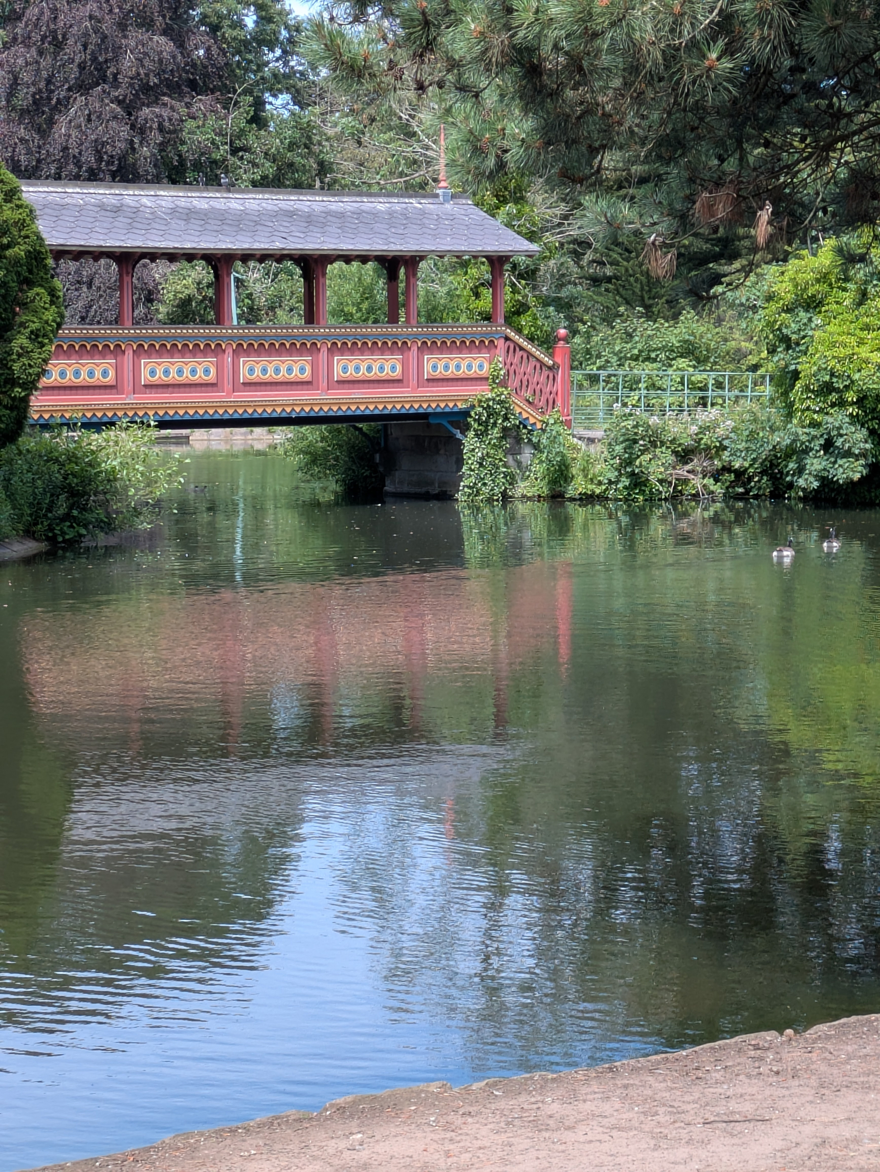A colorful covered bridge spans over a calm pond surrounded by lush green trees and foliage.