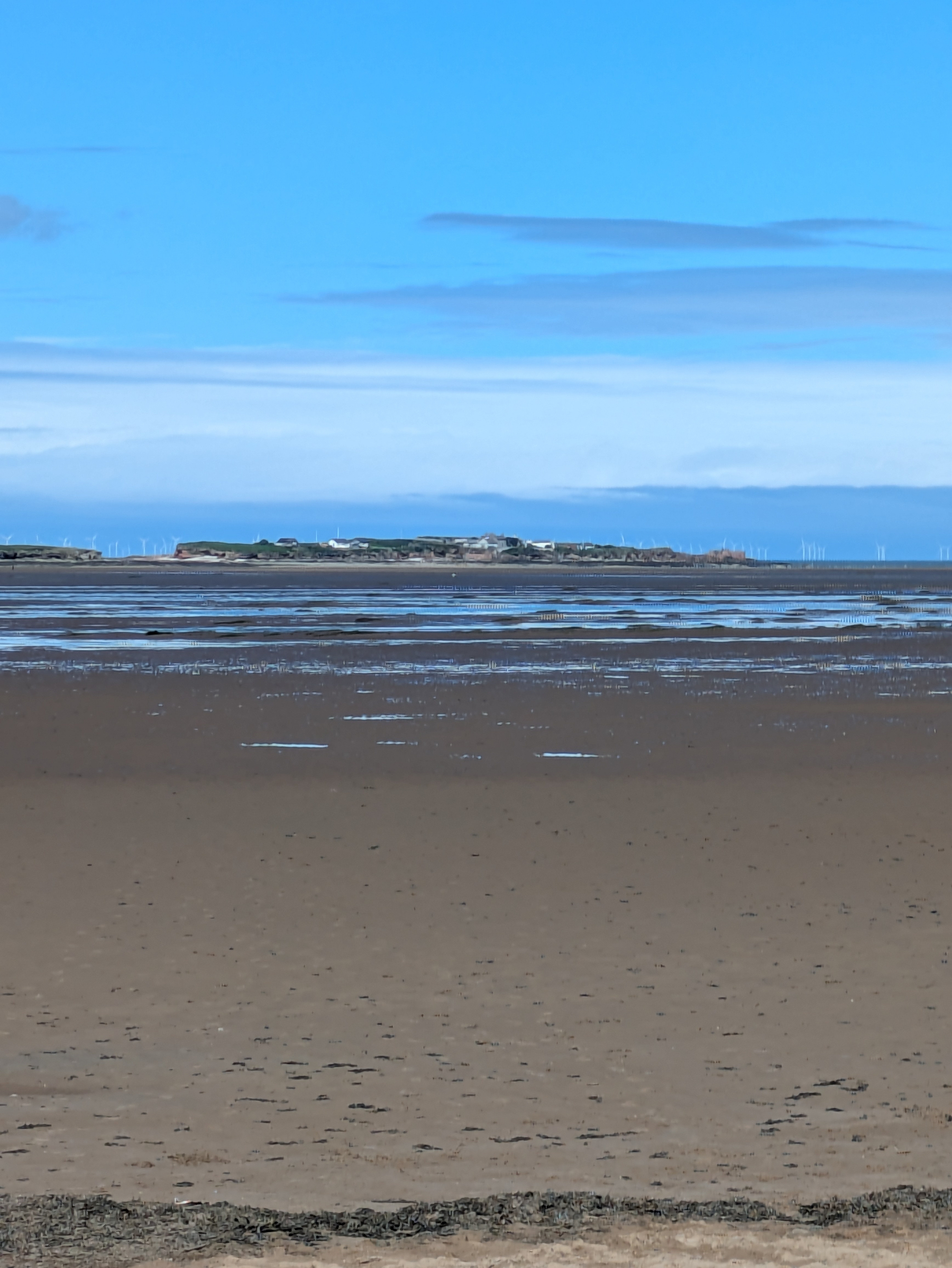 A sandy beach stretches towards a distant shoreline under a blue sky with scattered clouds.