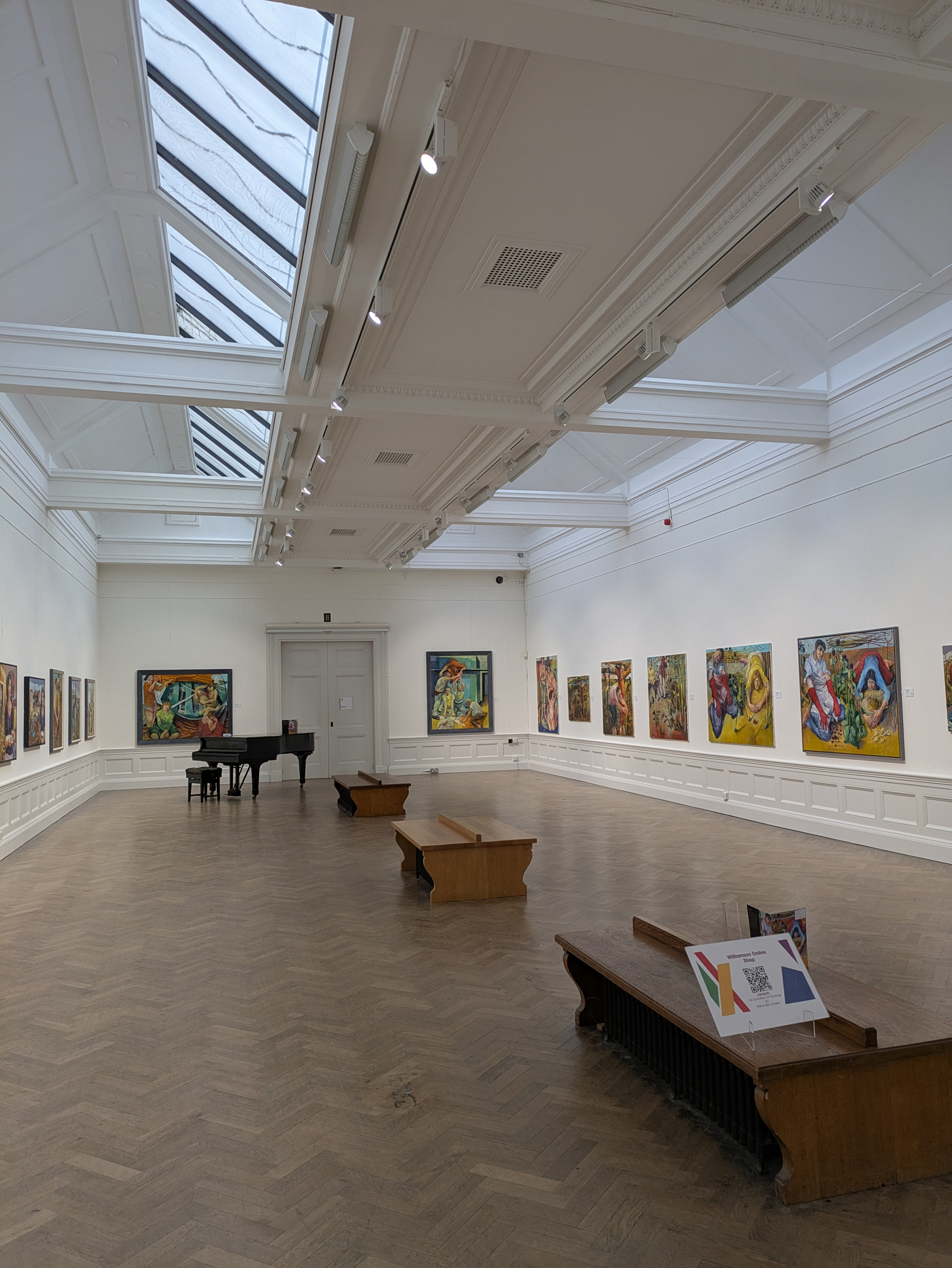 A spacious art gallery features colorful paintings on the walls, with benches and a piano placed in the center of the wooden-floored room.