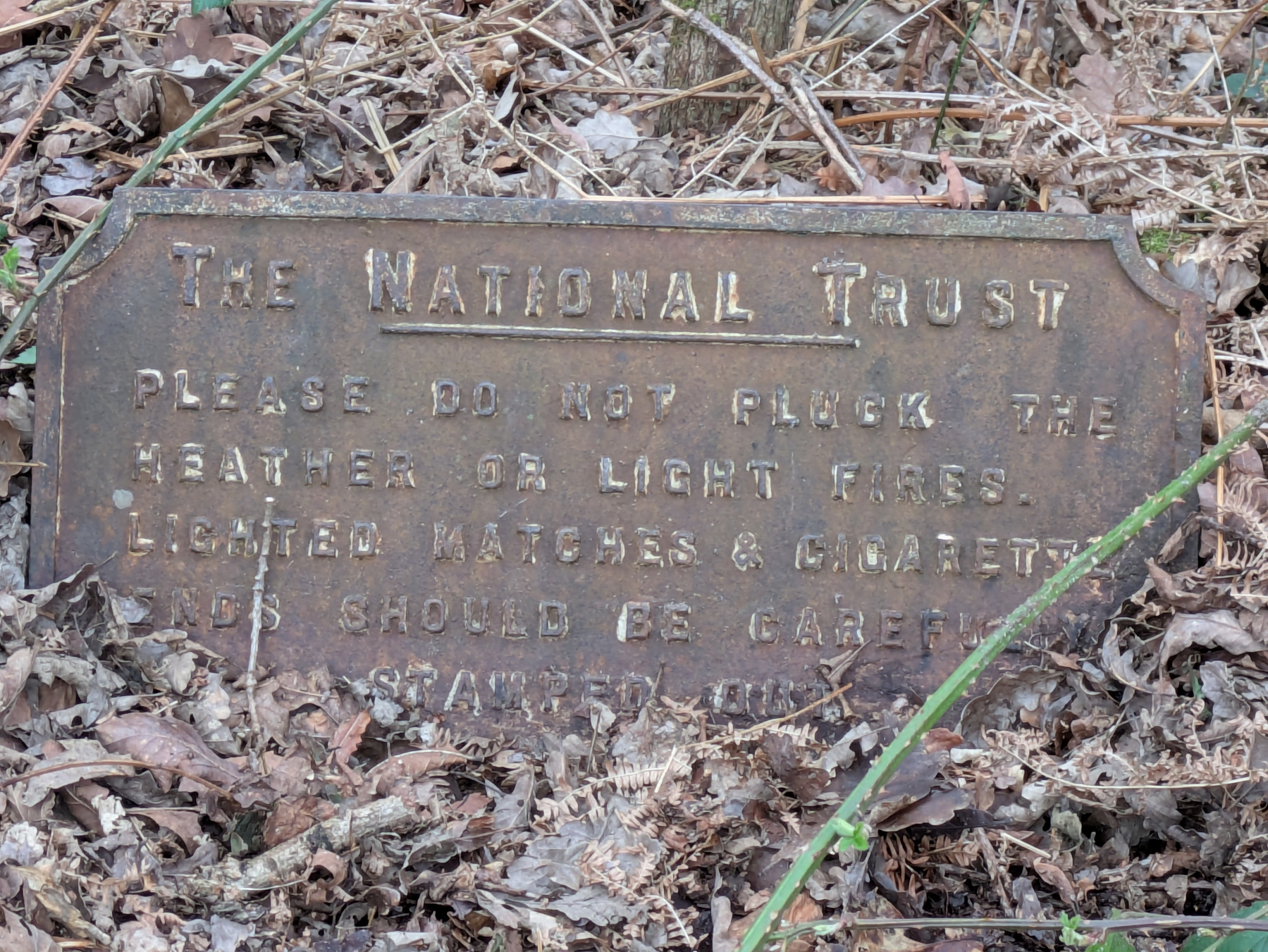 A weathered sign by The National Trust advises against picking heather or lighting fires in a wooded area.