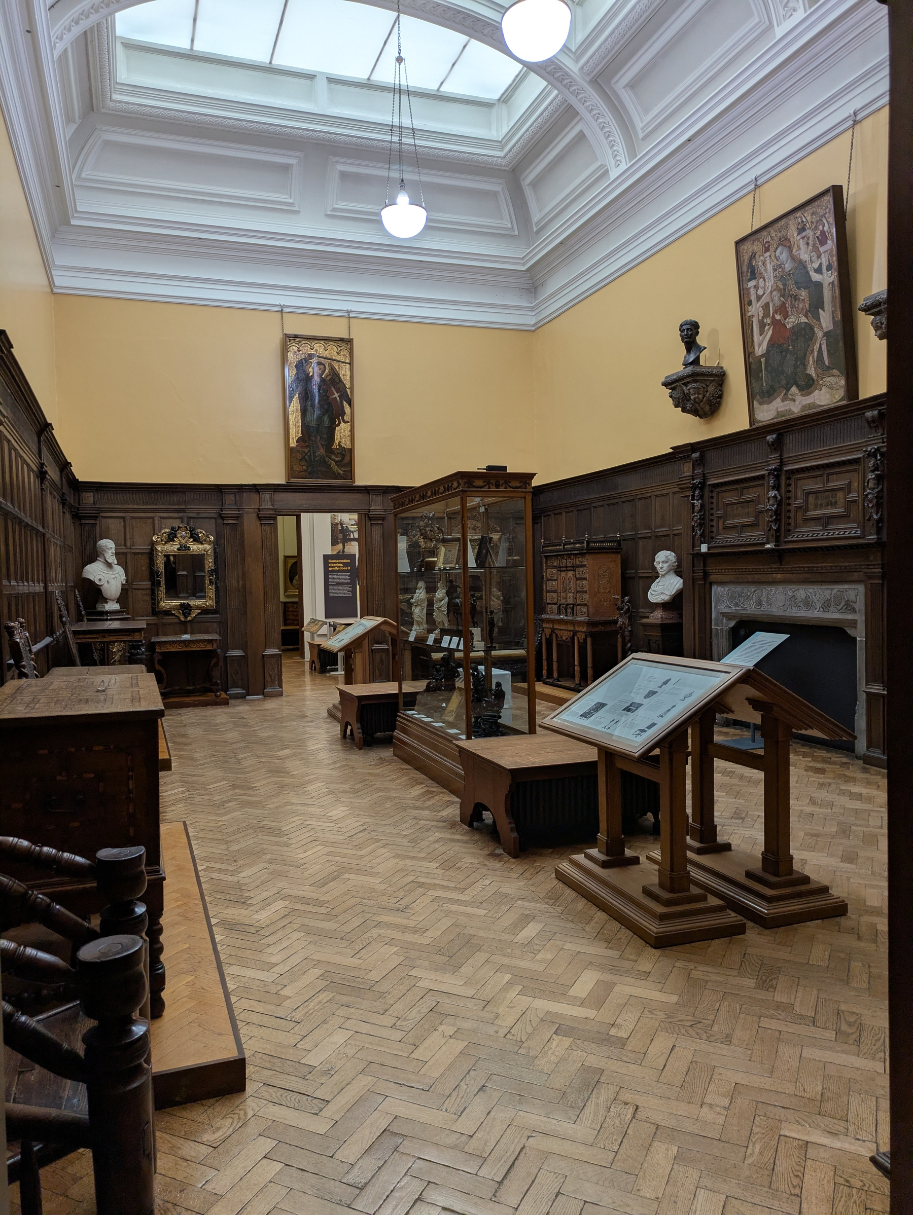 A museum gallery features busts, paintings, ornate furniture, and informational displays in a room with wood-paneled walls and a high ceiling.