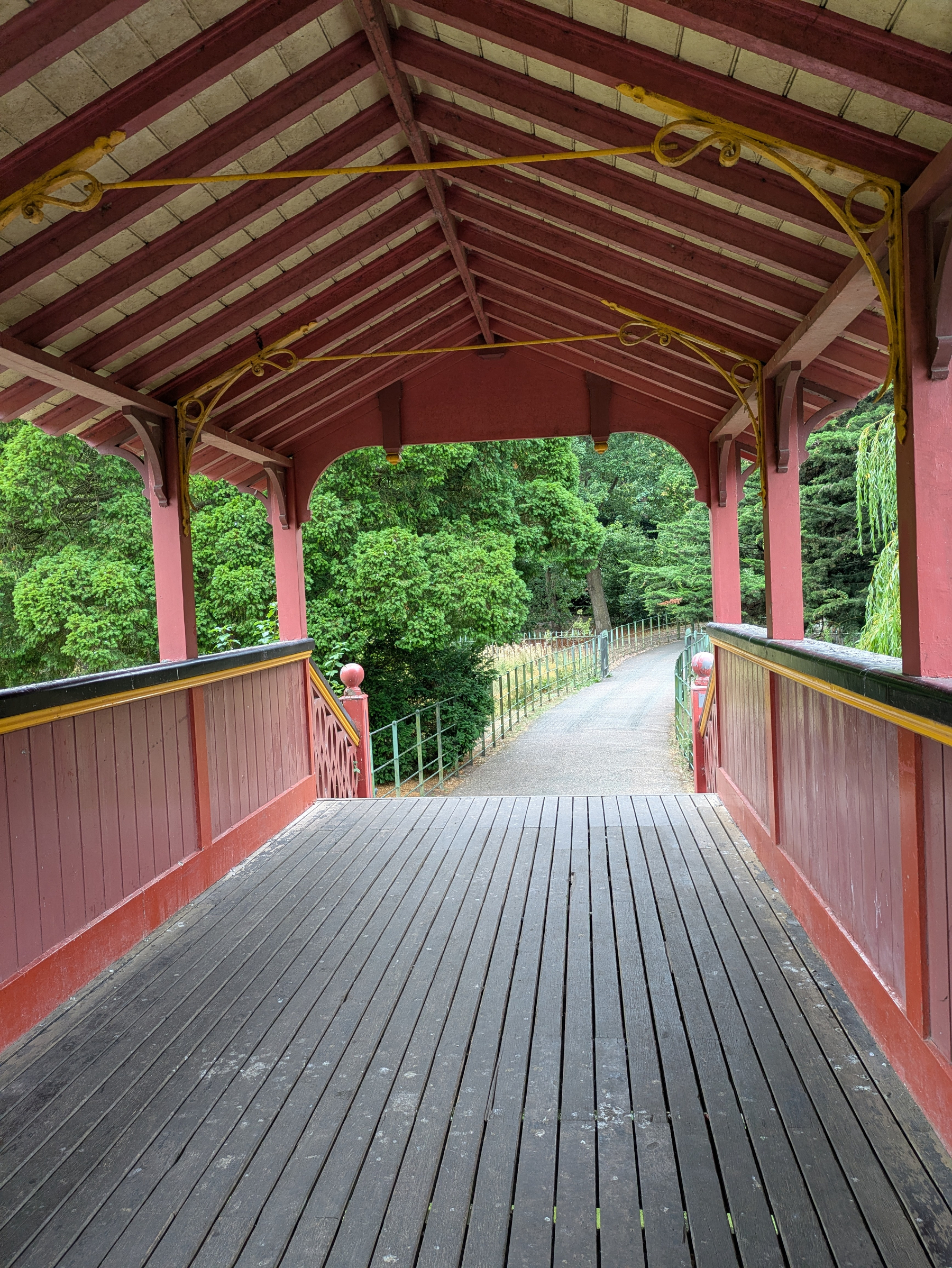 A wooden covered bridge painted red leads to a pathway surrounded by lush green trees.