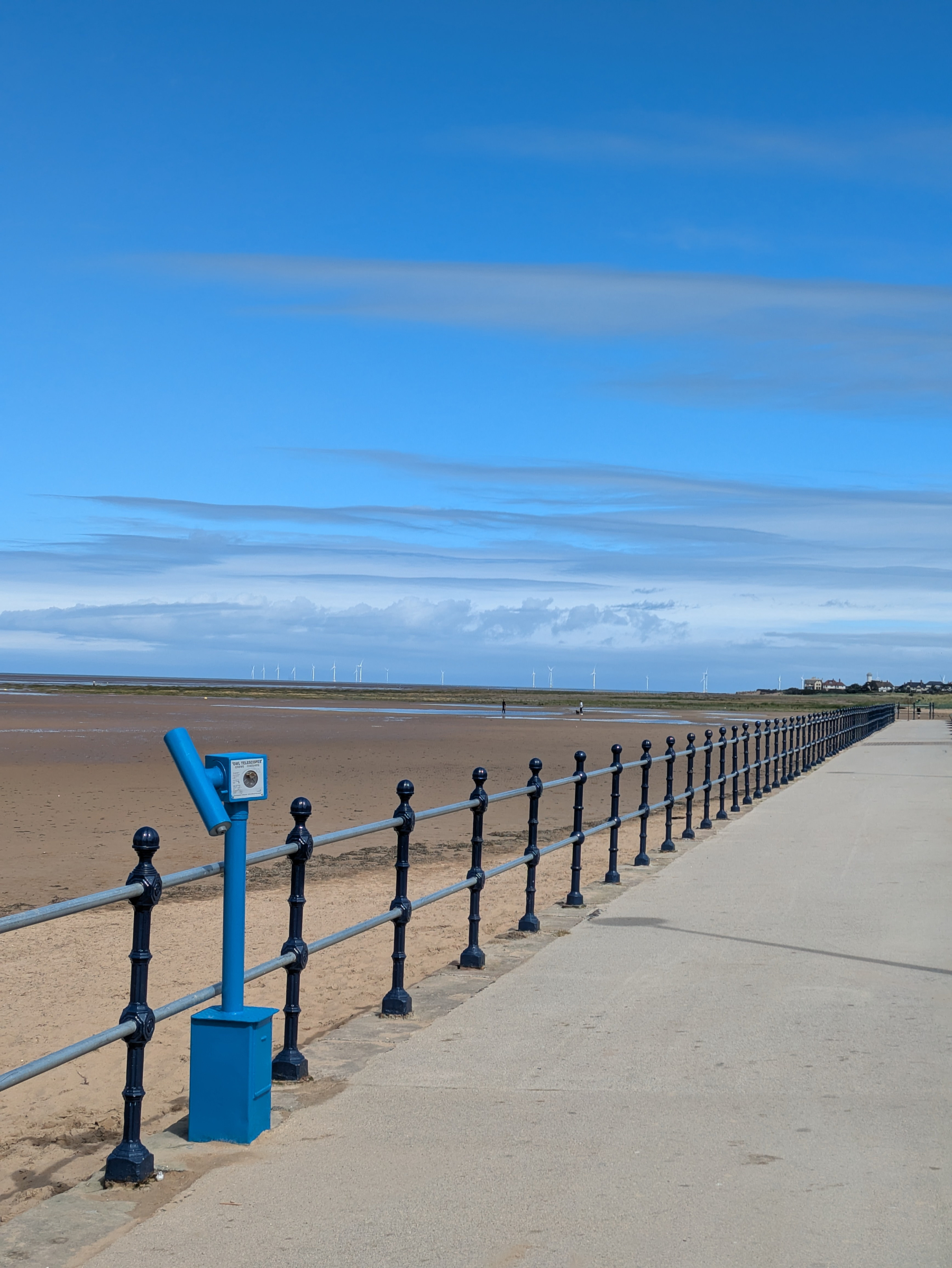 A seaside promenade features a long railing, a viewer telescope, and an expansive view of the beach and distant wind turbines under a blue sky.
