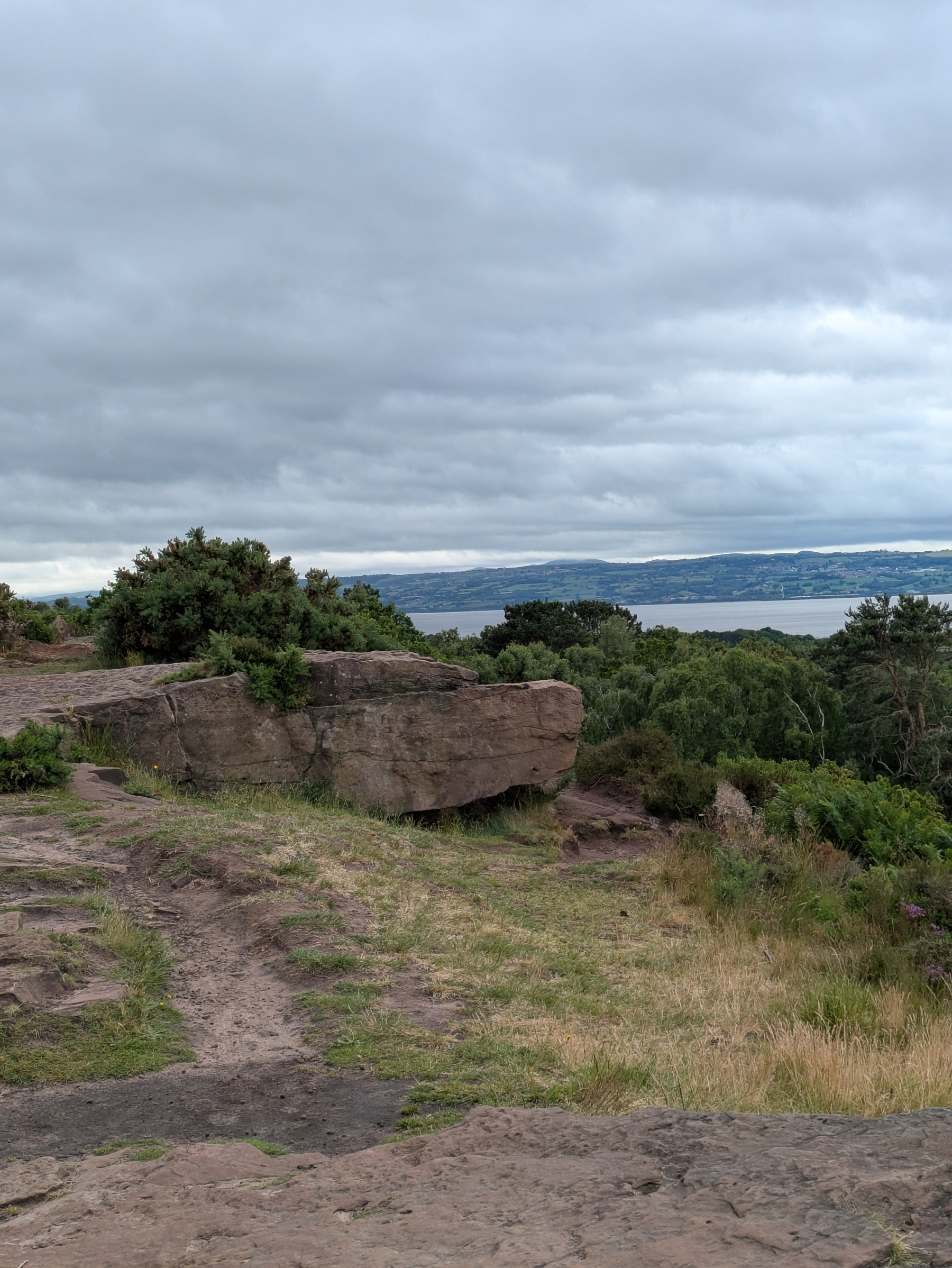 A rocky terrain with greenery overlooks a distant body of water under a cloudy sky.