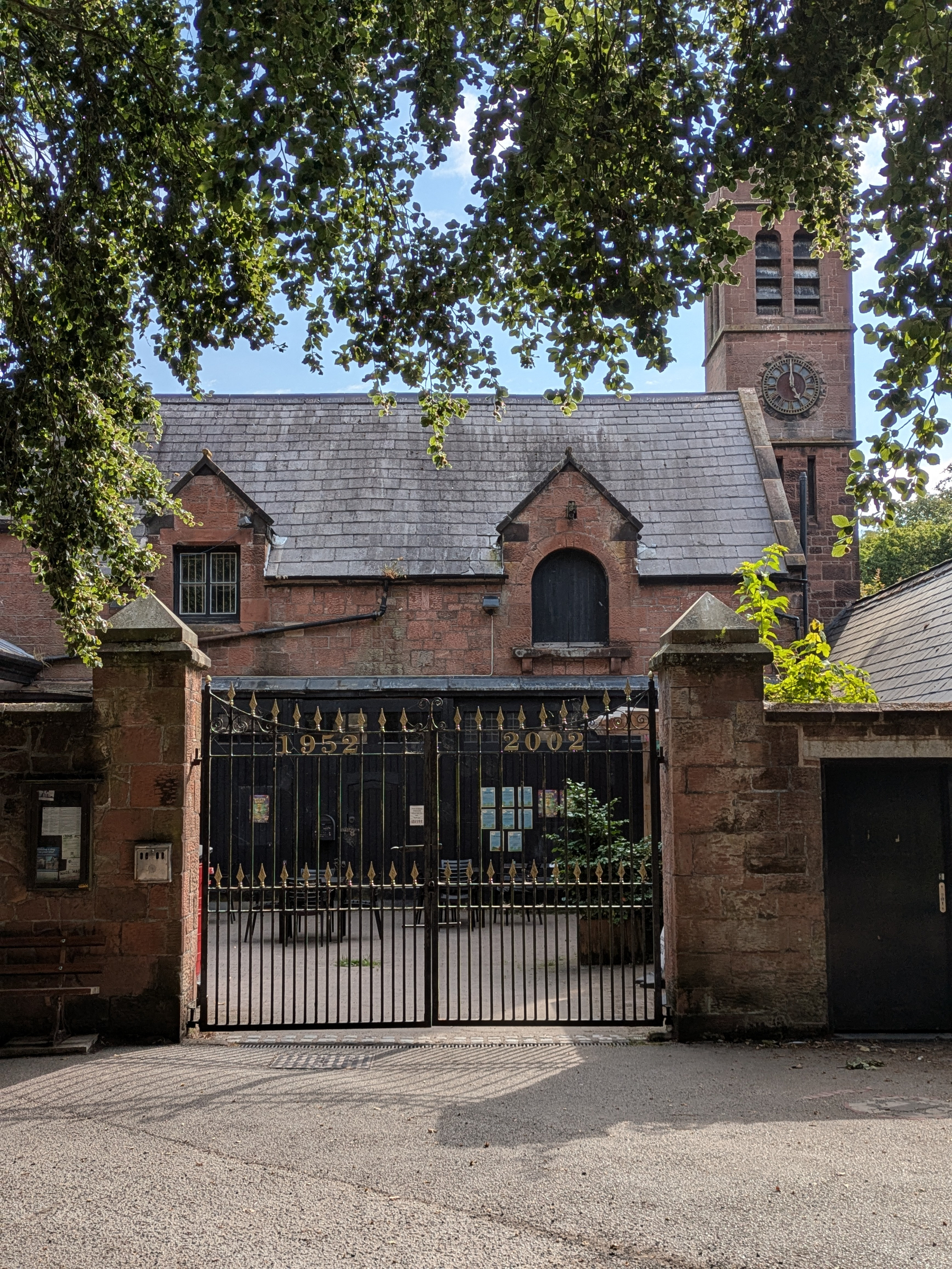 A historic brick building with a clock tower is seen behind a closed wrought iron gate, framed by leafy tree branches.